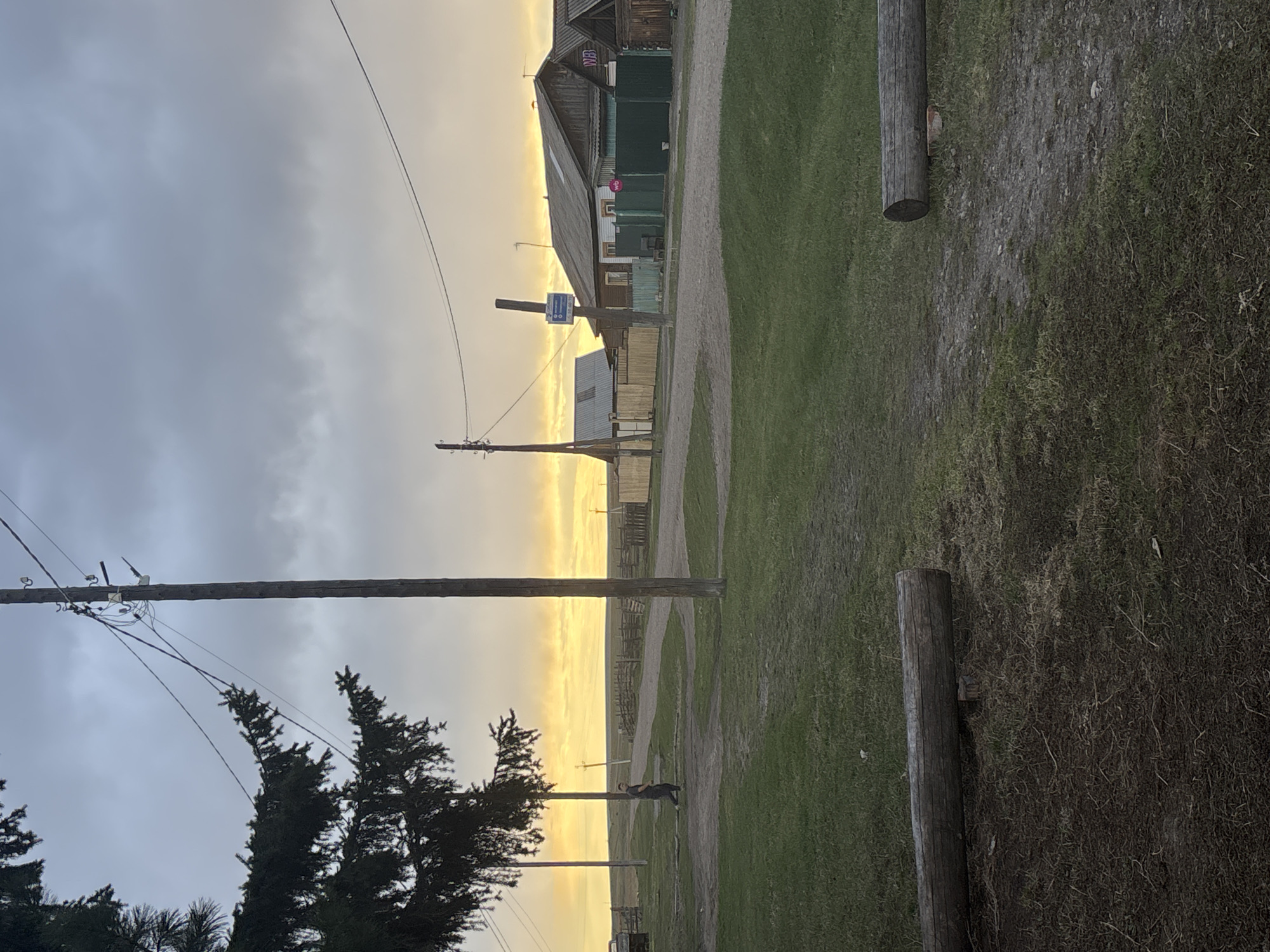 The image captures an outdoor scene in Zagatuy, Russia. It appears to be dusk or dawn, as the sky is a mix of gray clouds and golden light. A dirt road bisects the scene vertically, flanked by green grassy areas. Power lines and wooden poles are visible throughout. A distant figure of a person stands on the road. Buildings of wooden construction are also visible, with some signage in Russian. Wooden posts are in the immediate foreground on the sides of the dirt road.