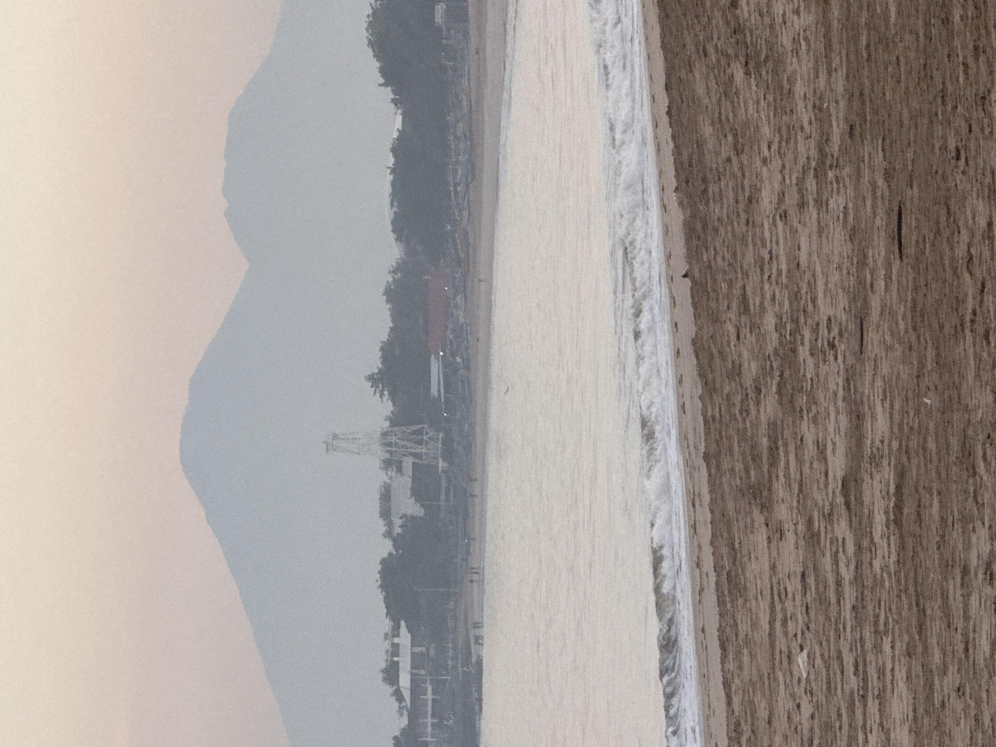 The image captures a serene beach scene in Kuta, Indonesia, with a focus on the natural elements and the subtle signs of human presence. The foreground is dominated by a sandy beach, showing the textured patterns of the sand, with a few small objects scattered on its surface. Beyond the sand, gentle waves with white foam roll towards the shore, creating a dynamic line where water meets land. The sea itself is a pale, reflective surface, almost shimmering in the diffused light.
In the middle ground, a shoreline is visible, lined with scattered trees and buildings. A notable structure, resembling a tall, skeletal tower or pylon, stands out against the hazy backdrop. Several small figures, appearing to be people, are spread out along the beach and near the water's edge, suggesting a calm, leisurely atmosphere. In the distance, the scene is framed by imposing, hazy mountains that rise against a soft, pastel-colored sky, indicative of either dawn or dusk. The overall impression is one of tranquility and natural beauty, with human activity minimally intruding upon the landscape.