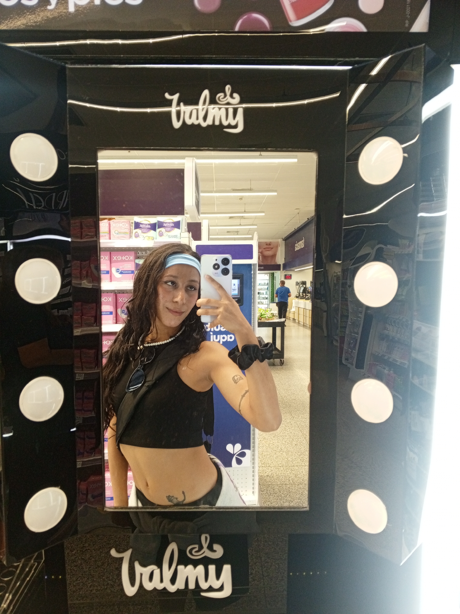 A young woman with long, wavy dark hair, a light blue headband, and a black crop top is taking a selfie in a makeup mirror. The mirror is black with "Valmy" written at the top and bottom, surrounded by white light bulbs. Her smartphone is in her right hand, angled upwards. She is wearing a pearl necklace and has a black scrunchie on her right wrist. A tattoo is visible on her right arm. The reflection in the mirror shows a store aisle filled with health and beauty products, including feminine hygiene products, and a man in a blue shirt standing near a checkout counter. The store's interior is brightly lit with fluorescent lighting.