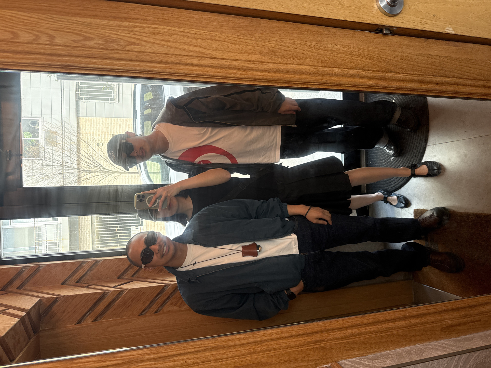 The image depicts a reflection in a storefront window, showing three individuals. The reflection reveals two men and one woman. One man wears a white t-shirt with a red target symbol, a jacket, a cap, and dark pants. Another man is wearing sunglasses, a white t-shirt, a blue jacket, and dark pants. The woman is wearing a black dress and holding a phone. The reflection also reveals a building across the street, a doormat, a door, and the wooden interior of the storefront where the reflection is captured. The buildings across the street appear to be in an urban area, which could be New York City.