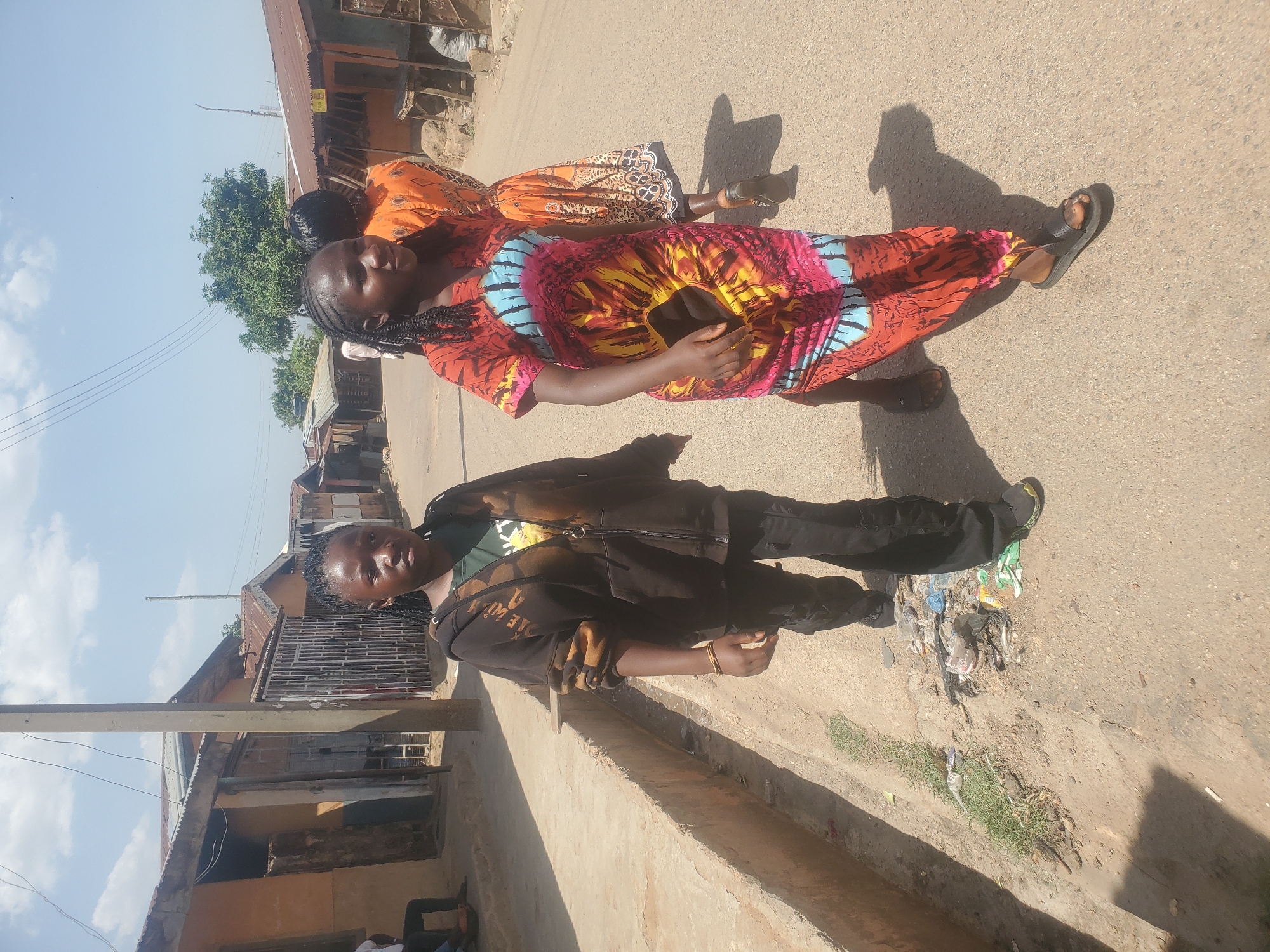 Two women stand side-by-side on a road in Jos, Nigeria. One woman wears a black jacket, black pants, and sneakers. The other wears a colorful, patterned dress and sandals. Behind them, a third woman is partially visible, wearing an orange patterned dress. A pile of litter is visible on the edge of the road near the woman in the black jacket. Buildings are visible on either side of the road. The sky is blue with scattered clouds. Overhead wires cross the sky.