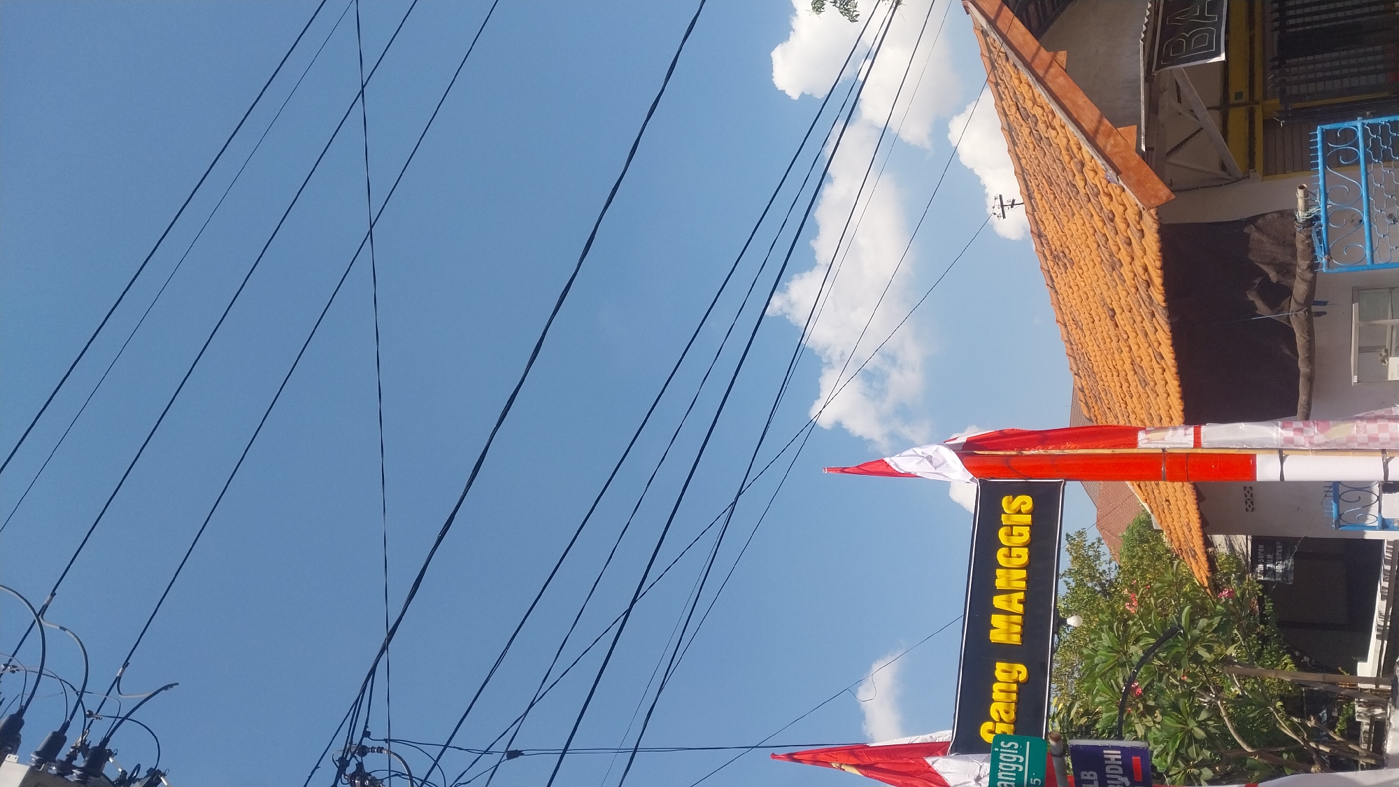 The image captures a street scene in Surabaya, Indonesia, under a bright blue sky with scattered white clouds. Prominently displayed is a sign reading "GANG MANGGIS" in large yellow lettering against a black background, indicating a street or alley name. Two Indonesian flags, red and white, are prominently featured, one attached to a pole next to the sign and another smaller one further to the left. The flags suggest a patriotic occasion or observance.
The foreground shows a section of a building with a terracotta-tiled roof and white walls. A dark brown awning covers part of the building's facade. Electrical wires are strung across the sky, a common sight in urban Indonesian settings. Below the sign, a smaller blue sign with white lettering is partially visible, with "LB DYUDHI" legible on it. The environment appears to be an ordinary street in a residential or commercial area, with trees and other buildings visible in the background. The overall impression is one of a typical day in a bustling Indonesian city, marked by local signage and national symbols.