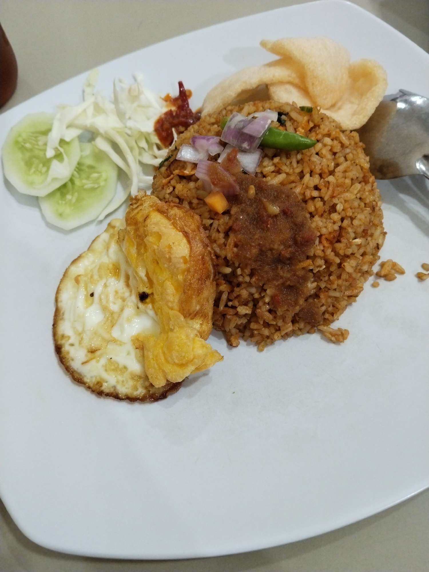 A plate of fried rice is presented on a white plate. The fried rice is topped with chopped red onions and green chilies. To the side of the fried rice, there is a fried egg with a runny yolk, sliced cucumbers, and shredded cabbage. Two light-colored crackers are perched atop the fried rice. A spoonful of chili paste is placed in the center of the fried rice. The scene is captured from a top-down perspective, with the plate resting on a neutral-colored surface. There is no indication of the specific location within Lhokseumawe, Indonesia.