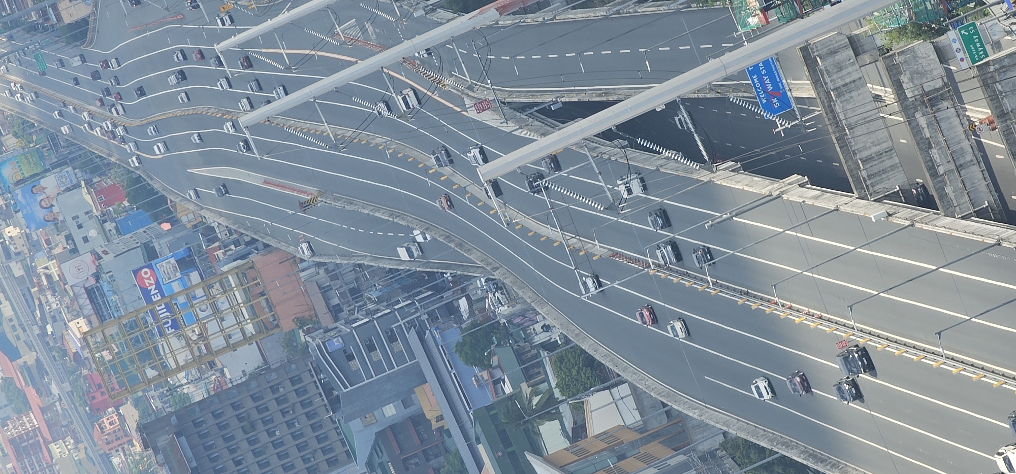 An aerial, high-angle, vertical, full shot captures a busy urban highway interchange in Makati, Philippines. The multi-layered road system, composed of concrete and asphalt, is filled with a variety of vehicles including cars, SUVs, and trucks, all moving in different directions. The highway is bordered by concrete barriers and illuminated by streetlights. Surrounding the highway are numerous buildings of varying heights and architectural styles, indicative of a dense urban environment. Some buildings are modern high-rises, while others appear older. Billboards, some featuring advertisements, are visible on some of the buildings. The overall scene suggests a typical day with heavy traffic flow within a bustling metropolitan area.