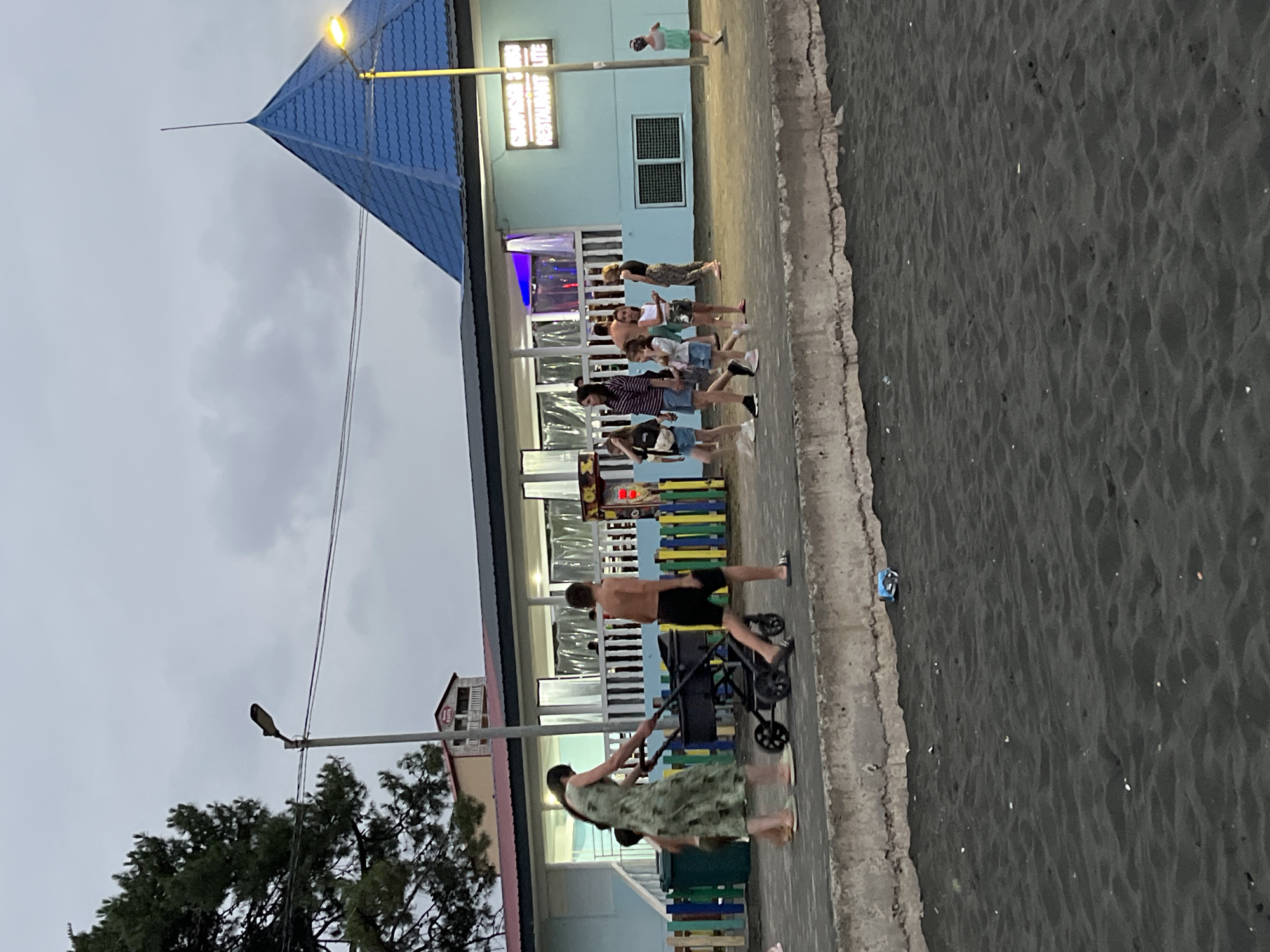 In Ureki, Georgia, on a beach with black sand, a group of people are walking along a concrete promenade. A woman in a green patterned dress is pushing a stroller. A young boy in black shorts walks beside her. To the right, several other people, including children, are walking. In the background, a light blue building with a blue, pyramid-shaped roof is visible. The building has windows and a sign that reads "GEORGIAN BREEZE RESTAURANT LITE". A street light is positioned near the building. The sky is overcast.