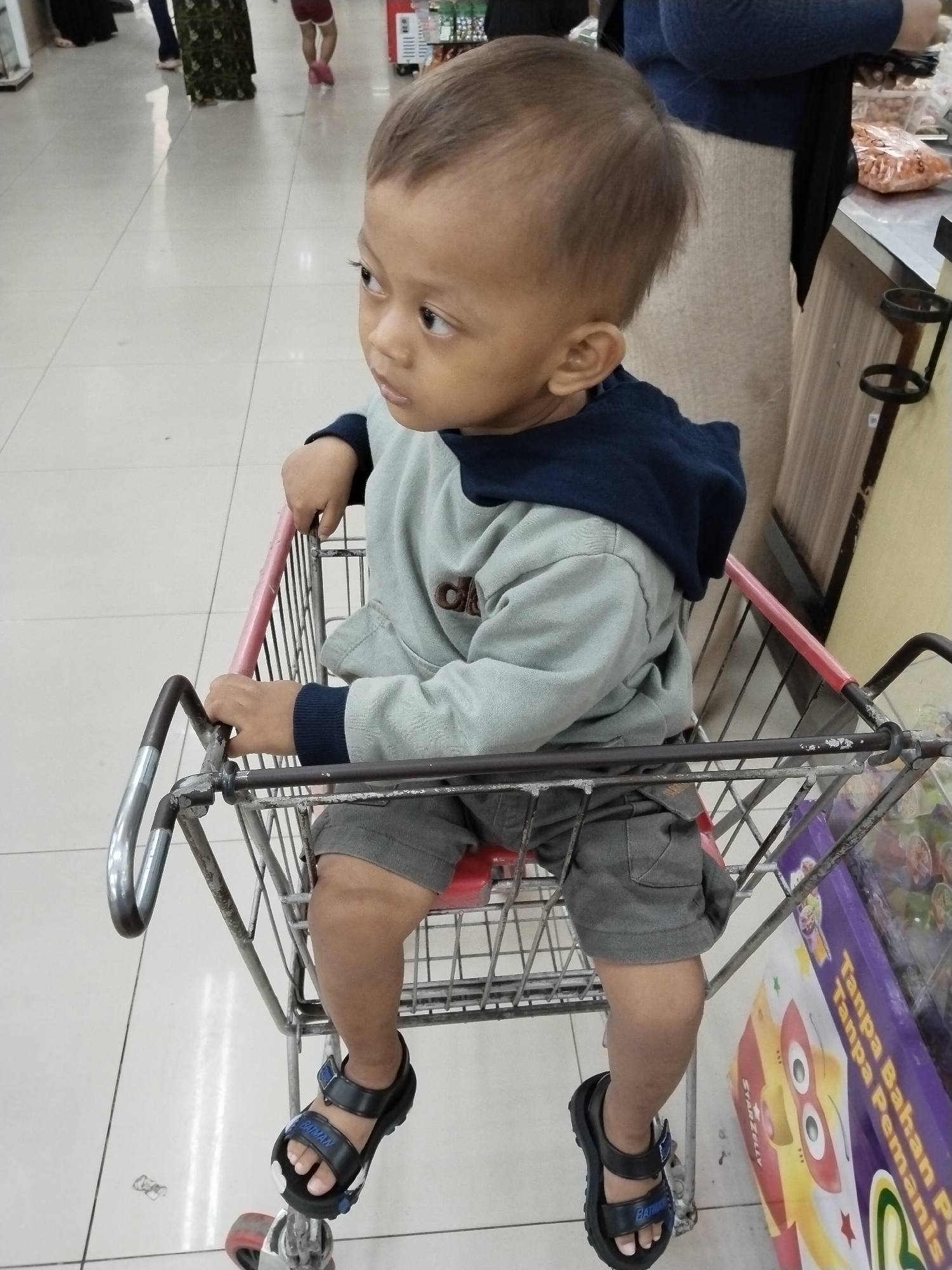 A young child, appearing to be between one and two years old, sits in a shopping cart in what is likely a grocery store in Kecamatan Pati, Indonesia. The child has dark hair and is looking to the side. They are wearing a light grey long-sleeved shirt with a dark blue hood, and grey shorts. The child is also wearing black sandals with blue accents. The shopping cart is made of metal and has a red handle. In the background, other shoppers are visible, along with shelves stocked with products. The floor is tiled. The lighting suggests an indoor environment.