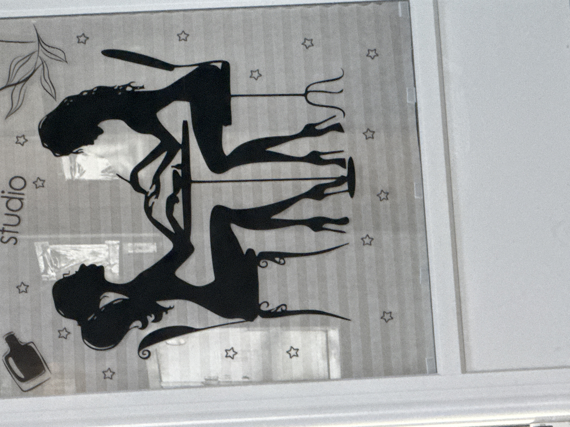 The image shows a window with a silhouette decal. The decal depicts two women sitting at a table, seemingly at a nail salon, with one woman filing the other's nails. There are stars and a nail polish bottle graphic also part of the decal. The text "studio" is visible above the women. The background of the window appears to be blinds. The location is not identifiable as Norilsk, Russia.