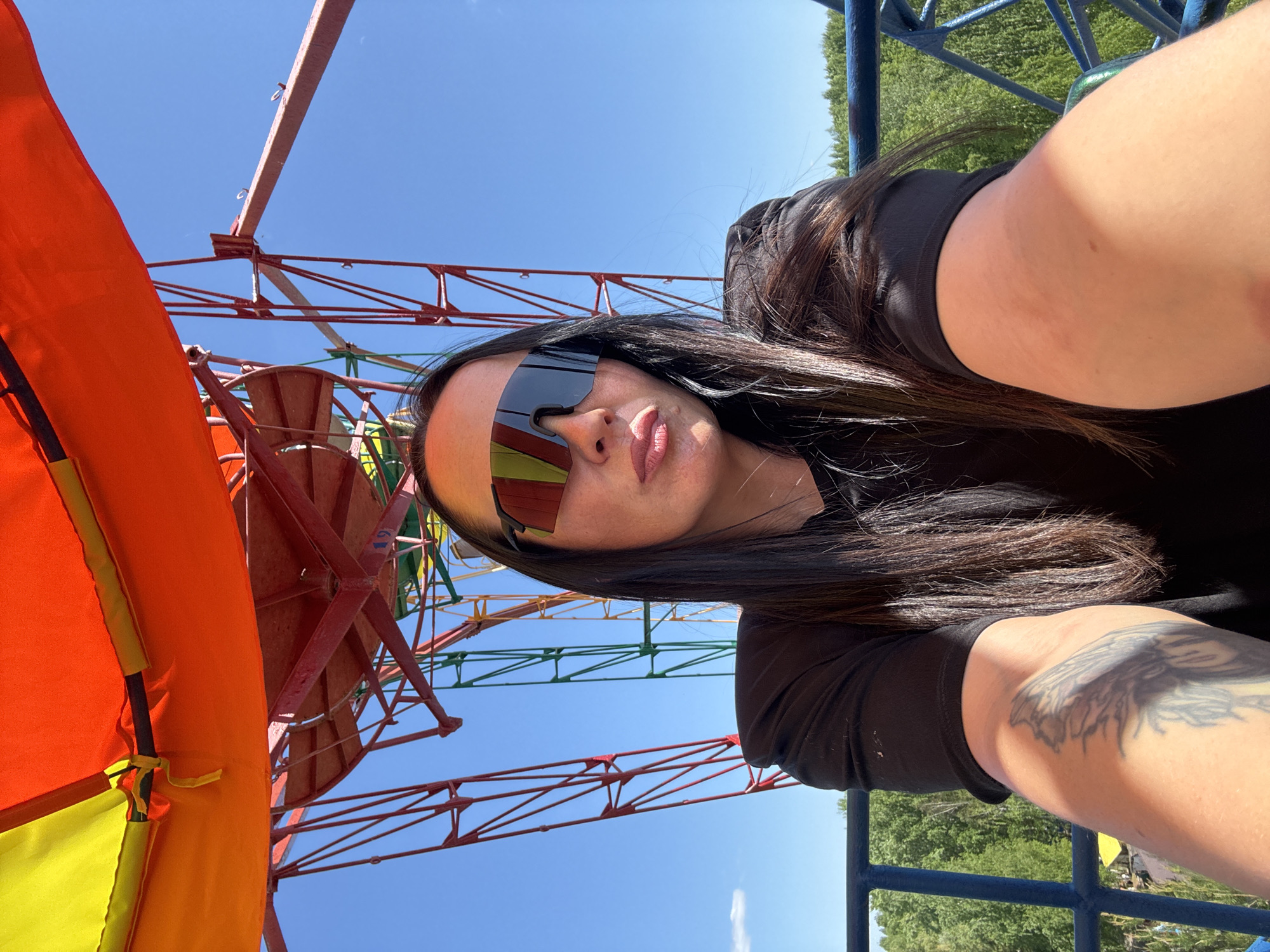 The image shows a woman with long dark hair wearing futuristic sunglasses. She is positioned in front of a Ferris wheel, with a colorful orange and yellow striped gondola visible on the left. The Ferris wheel's structure is primarily red, with some green and yellow sections. In the background, the sky is clear and blue. The woman has a tattoo on her arm and is wearing a black shirt. The setting appears to be Chaikovsky, Russia, based on the Ferris wheel and surrounding elements.