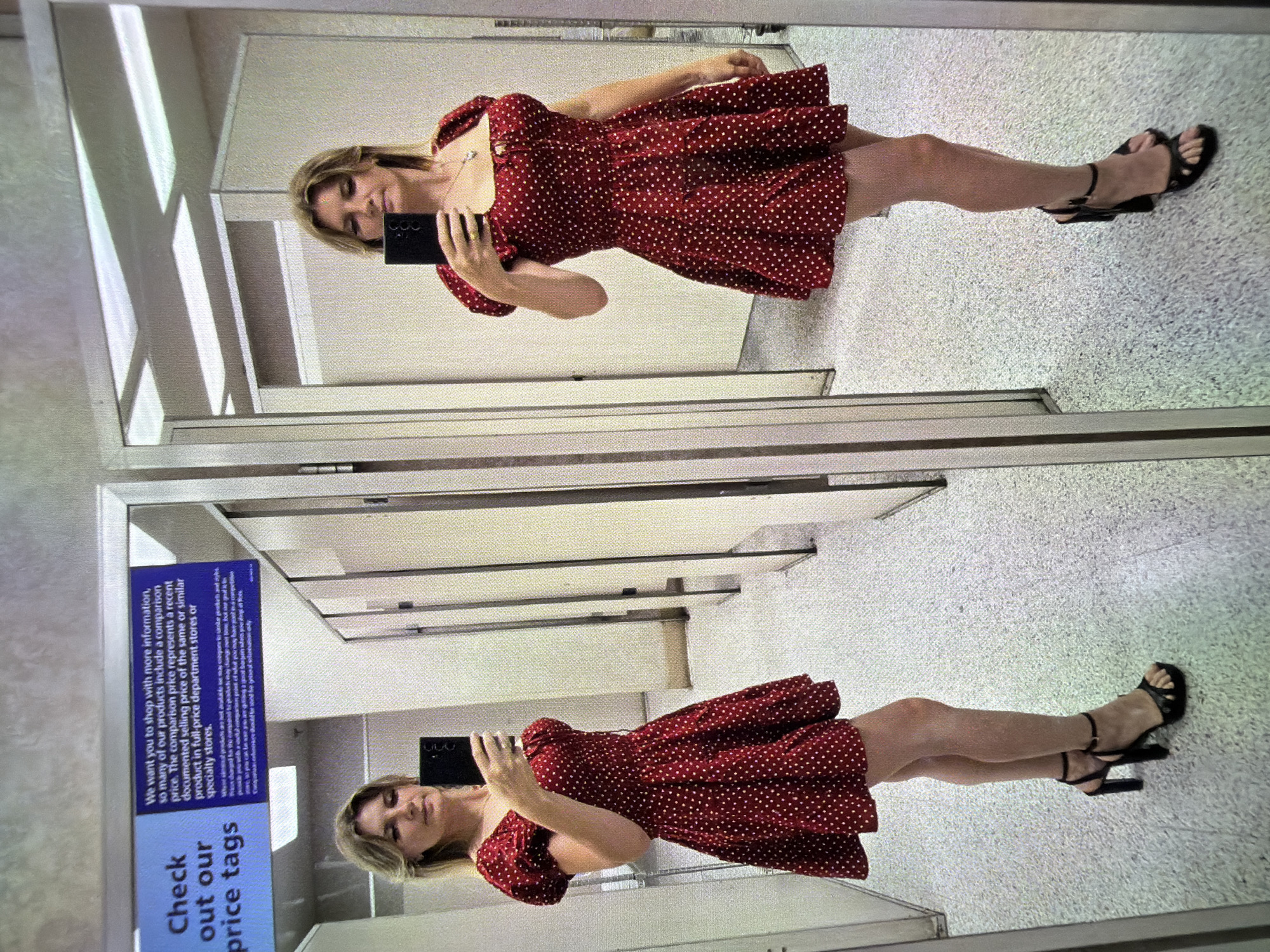 A woman is taking a selfie in a retail fitting room. She is wearing a short, red, polka dot dress with puffy sleeves and a square neckline. She is also wearing black strappy high-heeled sandals. The fitting room appears to be in a department store, with mirrors lining the walls. A sign in the background is visible, with text that reads "Check out our price tags." The floor is speckled linoleum, and the walls are paneled with vertical stripes. The lighting is overhead fluorescent. The woman's reflection is visible in the mirror, showing her from multiple angles. The scene is set in a fitting room, likely within a retail establishment. There are no visible indicators of the city being Sacramento, California, or the United States.