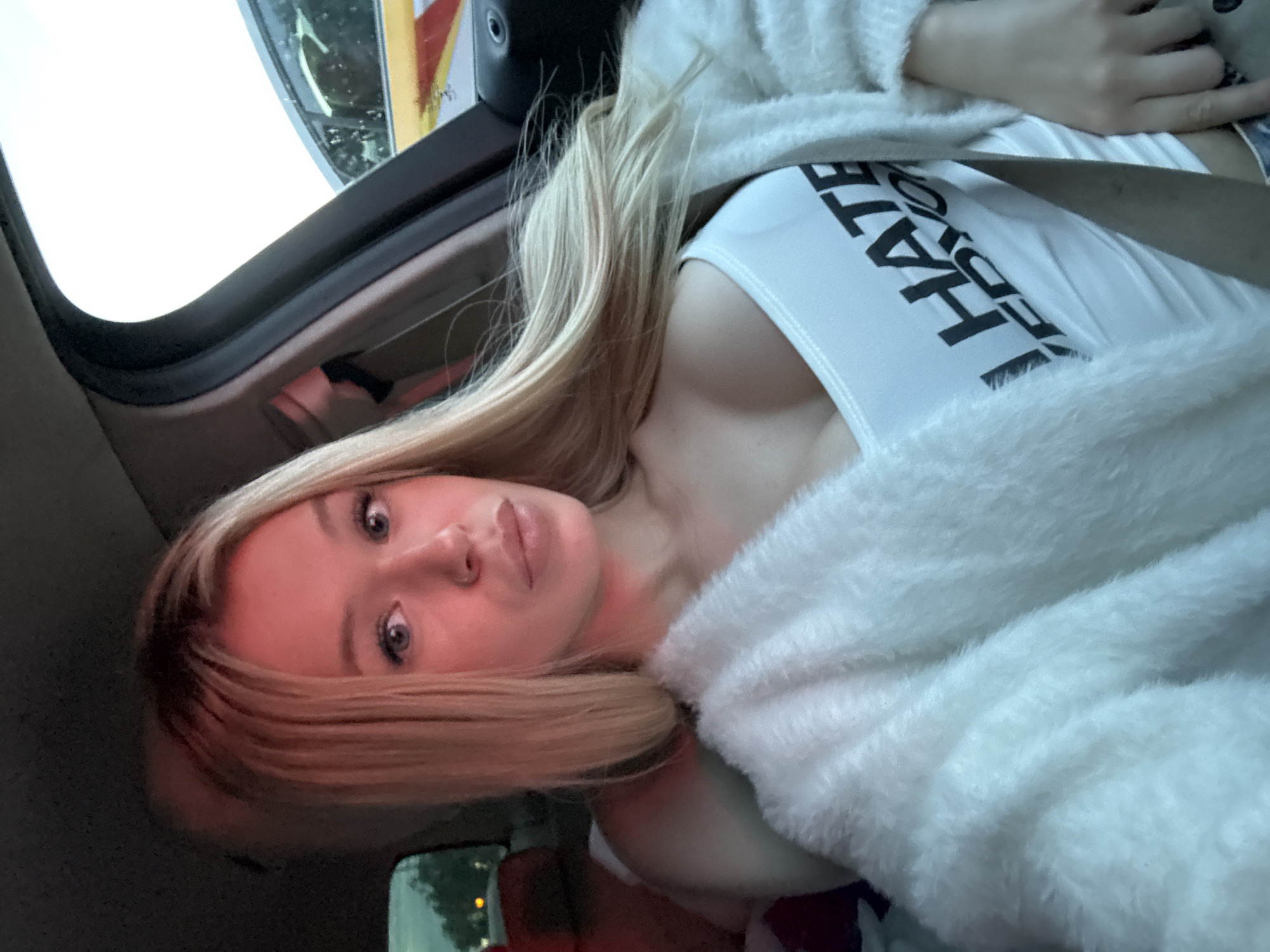 This is a portrait of a young woman with blonde hair, wearing a white tank top with black lettering that reads "I HATE YOU" and a white fuzzy sweater. She is inside a vehicle, possibly a car, with the door open to the outside. The lighting suggests it is daytime, and the scene appears to be outdoors, though the specific location is unclear. The woman is looking directly at the camera with a neutral expression. The overall mood of the image is candid and informal.