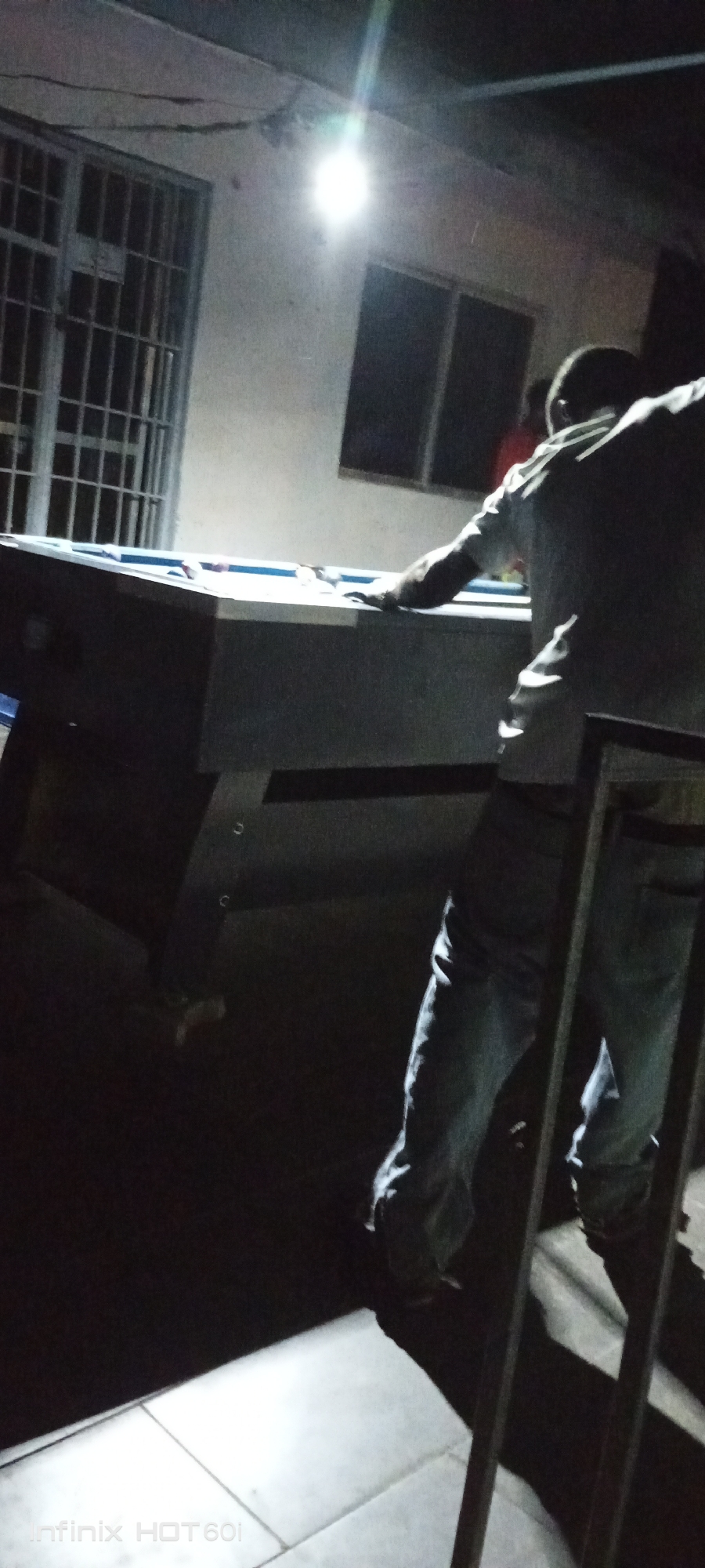 The image depicts a dimly lit scene, likely at night, in what appears to be an outdoor setting. A man stands at a pool table, his arm extended as if preparing to take a shot. He is wearing light-colored pants and a light-colored shirt. The pool table is positioned in front of a building that has a window with bars. An overhead light illuminates the area, casting strong shadows. Part of the frame of what might be a chair or table is visible in the foreground. A watermark 'Infinix HOT 601' is visible in the lower left corner, suggesting the device used to capture the image. The location is purported to be Adankolo, Nigeria.