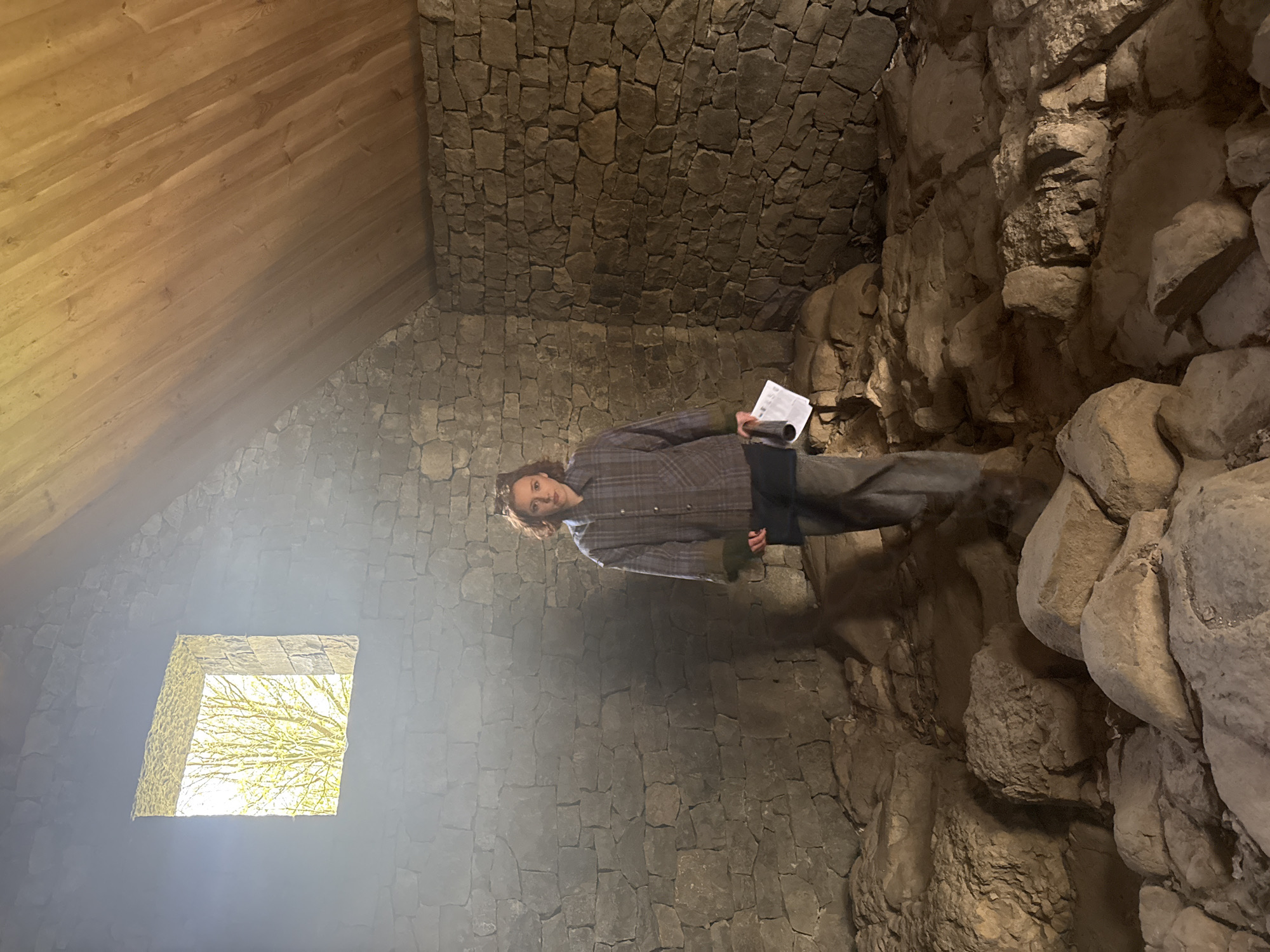 A medium, interior shot shows a person standing inside a stone structure, possibly a tower or old building. The person has shoulder-length brown hair and is wearing a plaid shirt and dark pants. They are holding a document or book in their hands. The walls of the structure are made of gray stones, and there is a small, square window on the left side of the frame through which light is entering. The ceiling appears to be made of wood. The scene is dimly lit. The location is unknown.
