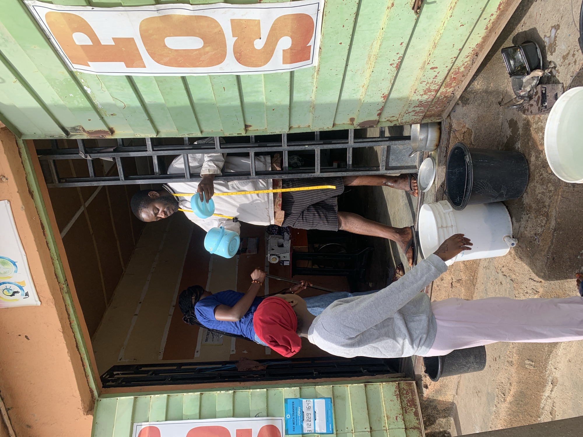 The image captures a bustling outdoor scene in Jos, Nigeria, during daylight hours, indicated by the bright sunlight and visible shadows. The setting appears to be the entrance of a small shop or service point, constructed from weathered green corrugated metal, with hints of rust. An orange-colored structure is visible to the left.
Three individuals are present. In the center, a dark-skinned man with a beard and short dark hair stands inside the green structure, facing left. He wears a light-colored long-sleeved shirt, striped shorts, and is barefoot on his visible foot. A yellow measuring tape is draped around his neck, and he holds a light blue scoop or small bowl in his left hand. Behind him and slightly to his left, a dark-skinned woman with braided hair is seated, wearing a blue top and blue jeans. She is focused on an object in front of her, which appears to be a sewing machine, indicating she is engaged in tailoring or similar work. To the right and closer to the camera, another person with lighter skin stands with their back mostly turned, wearing a grey hooded sweatshirt, light pink pants, and a red head covering. Their right hand rests on the rim of a white plastic bucket with a small spout.
On the ground in front of the green structure are several containers: a large black plastic bucket, the white plastic bucket mentioned previously, and another white bucket partially filled with water. A small metal pot and a black rectangular device (possibly a lamp or scale) are also on the ground.
Visible text includes a prominent white banner above the entrance with large orange letters that clearly show "OS" and part of a "P", likely spelling "POS" (Point of Sale). A smaller blue and white sign attached to the lower left of the green structure reads "PAY WITH TRANSFER", "NURTURE OPay", and displays a phone number "912 229 1643". The overall scene depicts a commercial or service exchange in an active community setting.