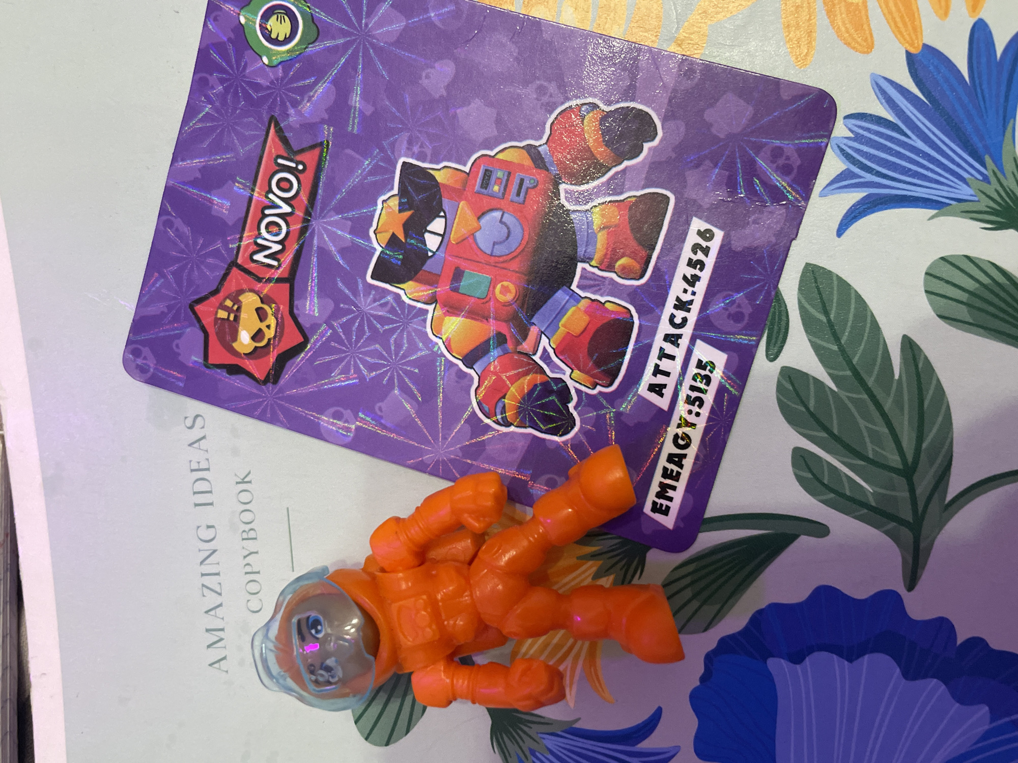 An overhead, slightly angled shot shows a collection of objects on a surface with a floral pattern. To the left, a small orange plastic toy figure, resembling an astronaut in an orange spacesuit with a clear helmet, stands upright. To the right, a purple holographic card, featuring a character that appears to be a robot with sunglasses and a star on its head, is positioned diagonally. The card has text labels including "NOVO!", "EMEAGY:5135", and "ATTACK:4526". The surface beneath the objects displays a white background with a repeating pattern of blue and green flowers and leaves. There is no indication or context related to Sinyavino, Russia in the image.