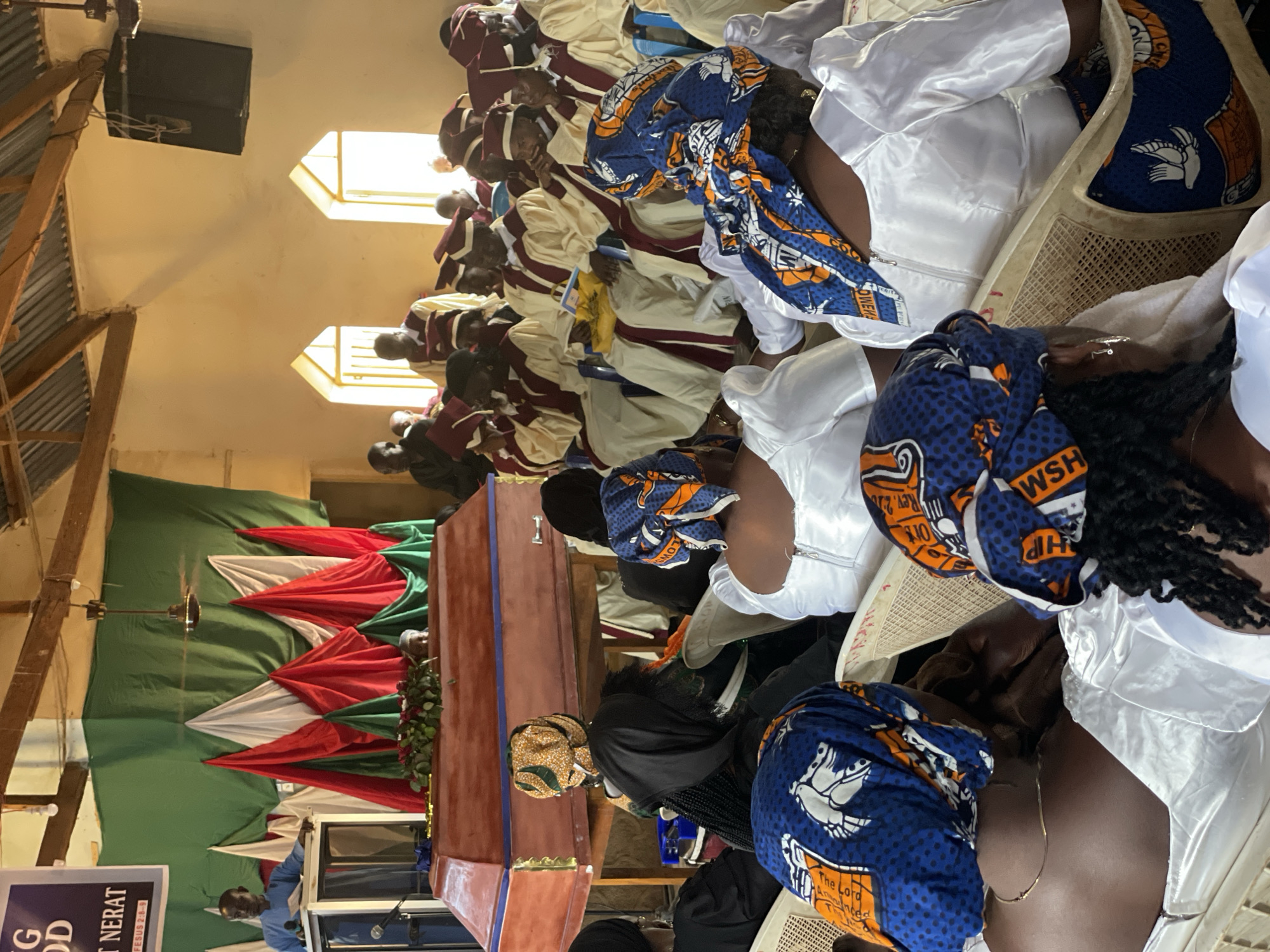 In Kassa, Nigeria, a funeral service is taking place inside a modest building that appears to be a church. The room is decorated with draped green, red, and white fabric. At the front of the room, a wooden coffin is positioned on a stand, adorned with a floral arrangement. A man in a blue shirt stands behind a podium, seemingly officiating the service. In the foreground and background, attendees are seated, many wearing white garments and colorful headwraps. Some individuals in the middle ground are dressed in ceremonial robes with maroon trim, suggesting a choir or official group. The environment is sparsely furnished, with visible wooden beams and corrugated metal roofing, indicating a simple structure. The lighting appears to be a mix of natural light from windows and overhead artificial lights.
