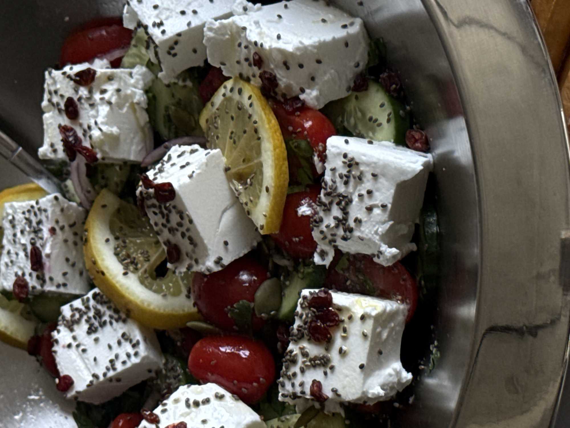 A close-up, high-angle shot shows a metallic bowl filled with a salad. The salad primarily consists of cubed feta cheese sprinkled with small, dark seeds. Slices of lemon, halved cherry tomatoes, and cucumber rounds are also present in the mix. Some red, dried berries are also seen. The bowl is likely made of stainless steel. No people are visible in the frame.
It's not possible to determine the location, including Yerevan, Armenia, from the visual information provided.