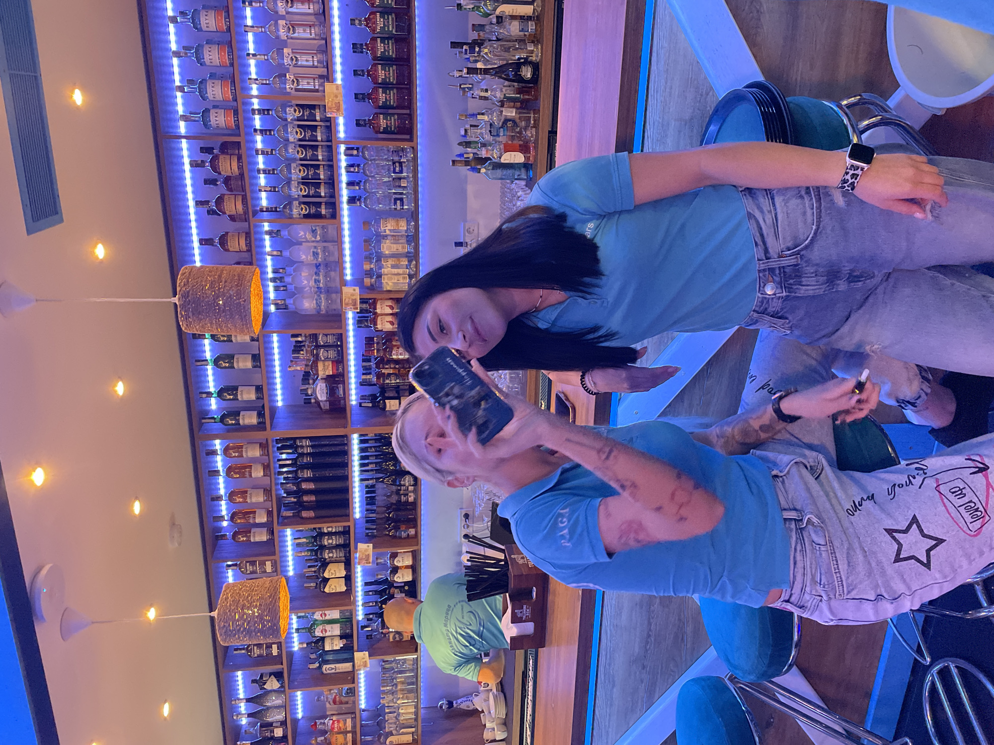 Two women are sitting at a bar. One woman with black hair is looking at the camera, while the other woman with blonde hair is holding a phone, presumably taking a picture. The bar has numerous shelves stocked with alcohol, illuminated by blue lights. There are pendant lamps hanging from the ceiling. The woman taking the picture is wearing jeans with a star and the words "Level Up" printed on them. The bar appears to be located in Plovdiv, Bulgaria, based on the Cyrillic inscription "Блямата Марица."