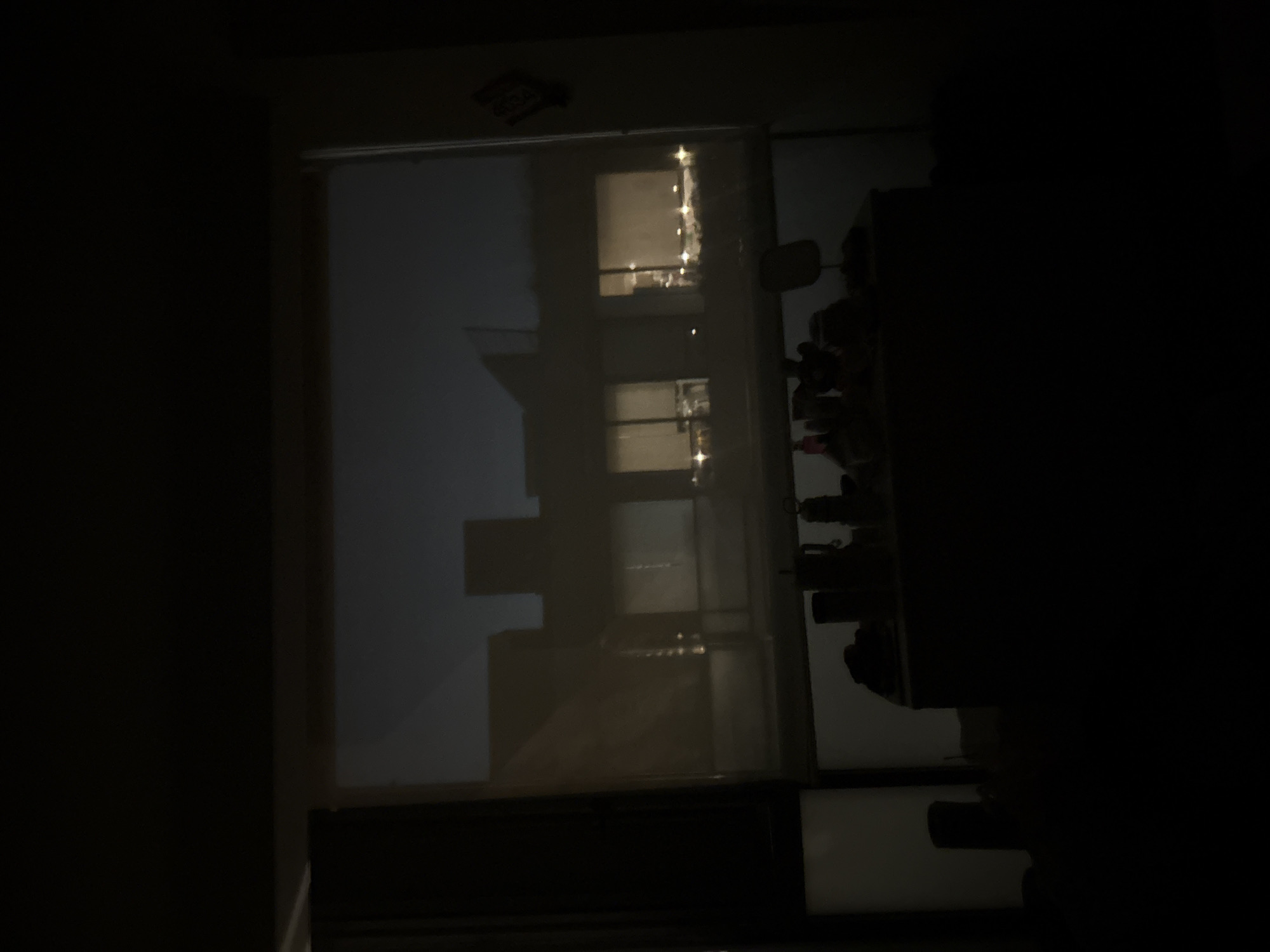 The image shows a dimly lit room at night. Through a window with a partially drawn blind, there is a reflection or view of buildings across the way, possibly in Mexico City based on the prompt. The scene is dark, obscuring details of the room's contents, but one can make out a desk or surface with items on it against the right side of the window. The overall atmosphere is one of darkness and limited visibility.