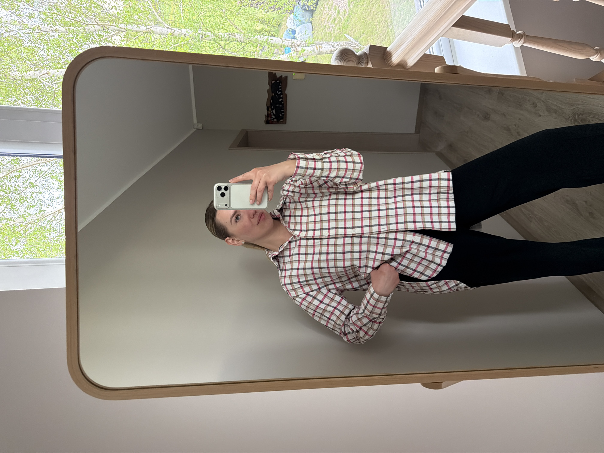 A woman is taking a picture of herself in a mirror inside a room. She is wearing a plaid shirt and black pants. The mirror reflects a hallway with stairs. Outside the window is green foliage. The location is suggested to be Dreiliņi, Latvia.