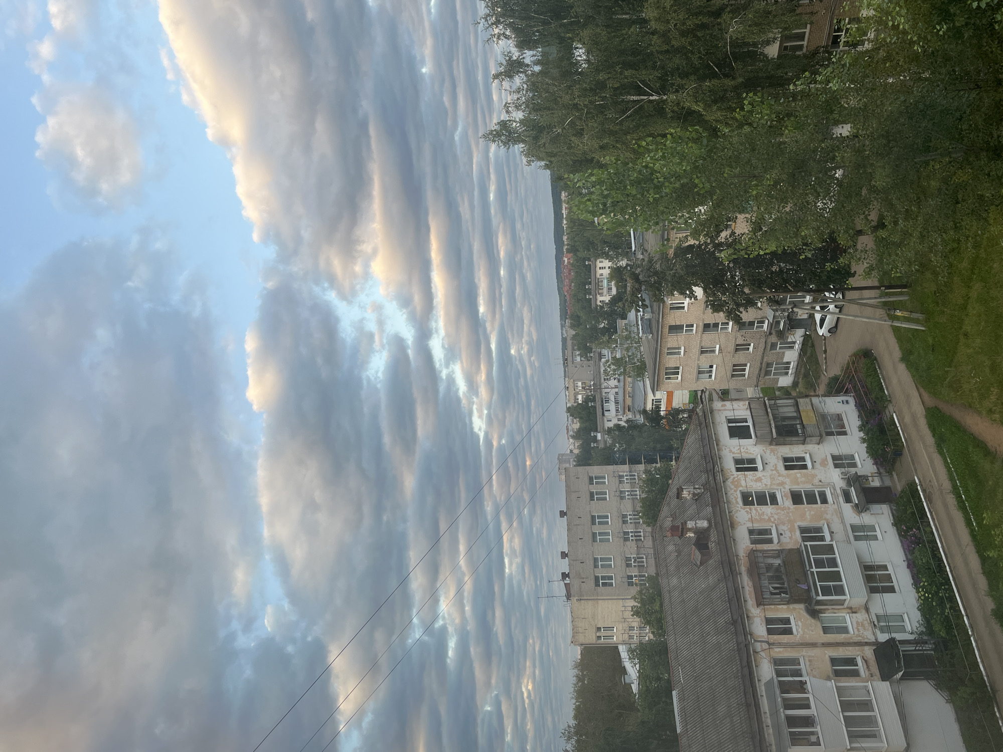 The image depicts a view of a residential area in Chaikovsky, Russia, under a cloudy sky. The foreground features the roof of a weathered, multi-story apartment building with a gray, corrugated metal roof. Several windows, some with balconies, are visible on the building's facade, which is painted in pale, peeling colors. Below the building, a paved road curves around it, with patches of green grass and flower beds along the edge.
In the mid-ground, more apartment buildings of varying heights and architectural styles are scattered amongst lush green trees. Some of these buildings are brick, while others are painted. A white car is parked on the road in front of one of the buildings. Power lines are strung across the scene.
The background reveals a dense forest canopy and the distant outlines of more buildings, suggesting a suburban or semi-urban landscape. The sky is filled with a mix of gray and white clouds, with pockets of pale blue sky visible. The lighting suggests it is either early morning or late afternoon, with the sun casting a soft glow on some of the clouds. No people are visible in the image.