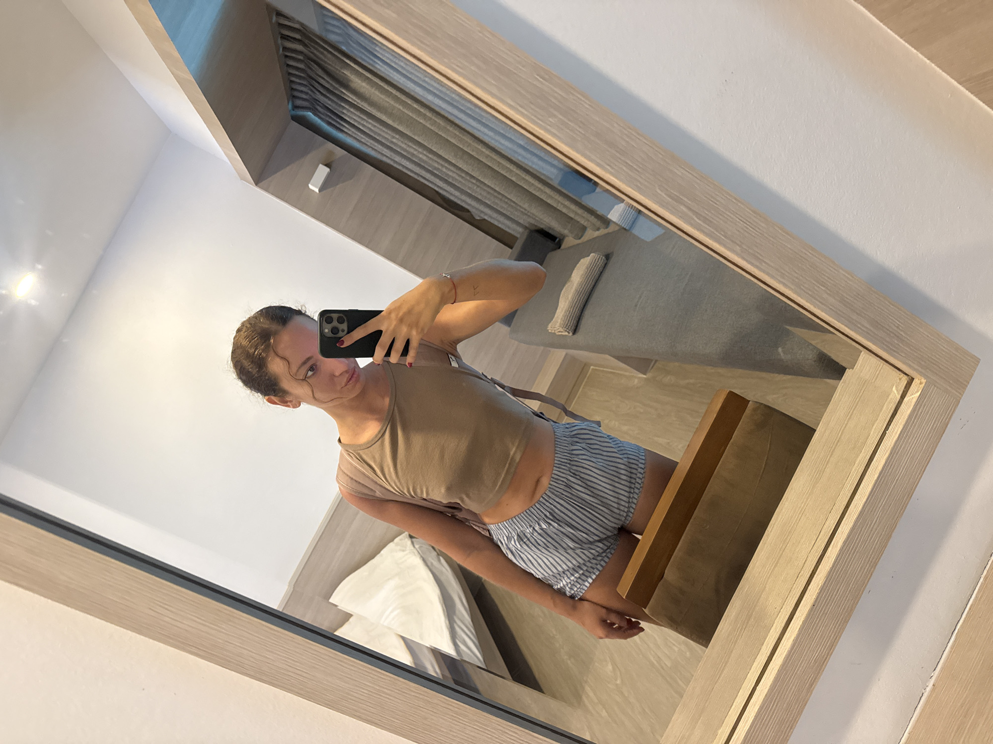 A young woman, with her hair pulled back in a bun and wearing a beige crop top and blue and white striped shorts, takes a mirror selfie in what appears to be a hotel room. She is holding a black smartphone in her right hand. In the reflection, a bed with white linens is visible behind her. To the right, a gray sofa with a folded gray towel on it can be seen, along with dark gray curtains. A wooden framed mirror dominates the foreground. The floor is a light wood laminate. A brown paper bag is placed on a wooden object in the lower left corner of the frame. The lighting suggests it is daytime. The location is specified as Ko Samui District, Thailand.
