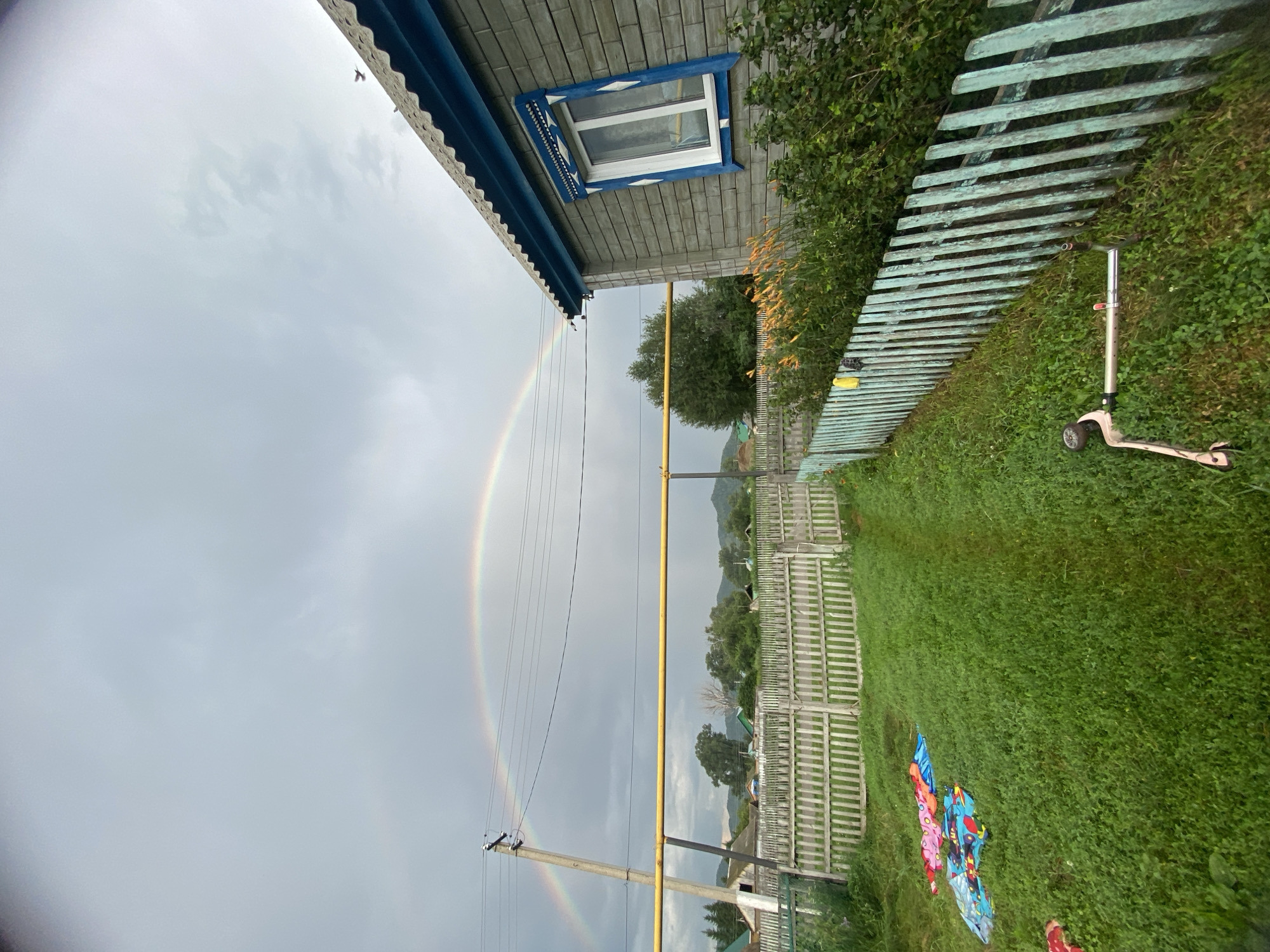 The image depicts an outdoor scene in the village of Maloarslangulovo, Russia, under a cloudy sky with a rainbow arching across it. On the right, a gray brick house with a blue-trimmed window is visible. A light blue wooden fence runs along the foreground, separating the yard from the street. The yard is covered in green grass, with a pathway leading through it. Colorful blankets are spread on the grass near the fence. In the background, more houses, a wooden fence, and trees are visible, with hills in the distance. A utility pole with wires and a yellow pipe are present on the left side of the image. A scooter is lying on the grass near the fence on the right side of the image.
