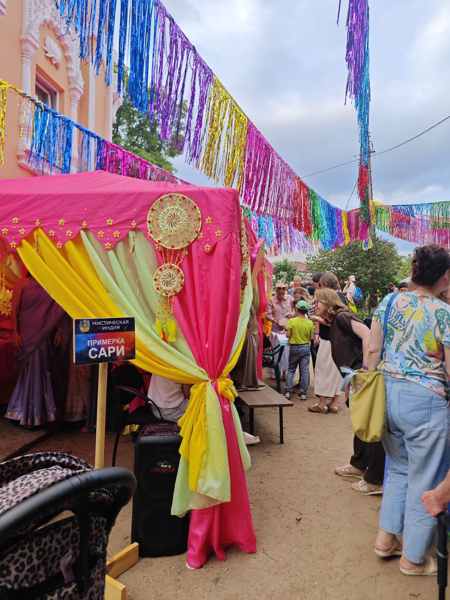 This image captures an outdoor event, likely a festival or cultural gathering, in Saint Petersburg, Russia. The scene is adorned with vibrant, multicolored metallic streamers hanging from poles and across the overhead space, creating a festive atmosphere. In the foreground, a pink tent-like structure is draped with bright yellow and green fabrics. A sign attached to a wooden pole reads "МИСТИЧЕСКАЯ ИНДИЯ" (Mystical India) and "ПРИМЕРКА САРИ" (Sari Fitting), indicating a theme related to Indian culture and the opportunity to try on saris. Several people are present, some interacting with the tent, others observing or conversing. A black speaker is placed near the tent, suggesting music or announcements are part of the event. The ground appears to be a dirt or gravel pathway, and the background shows trees and a cloudy sky. The overall impression is of a lively and colorful public event taking place in an urban or semi-urban outdoor setting in Saint Petersburg.