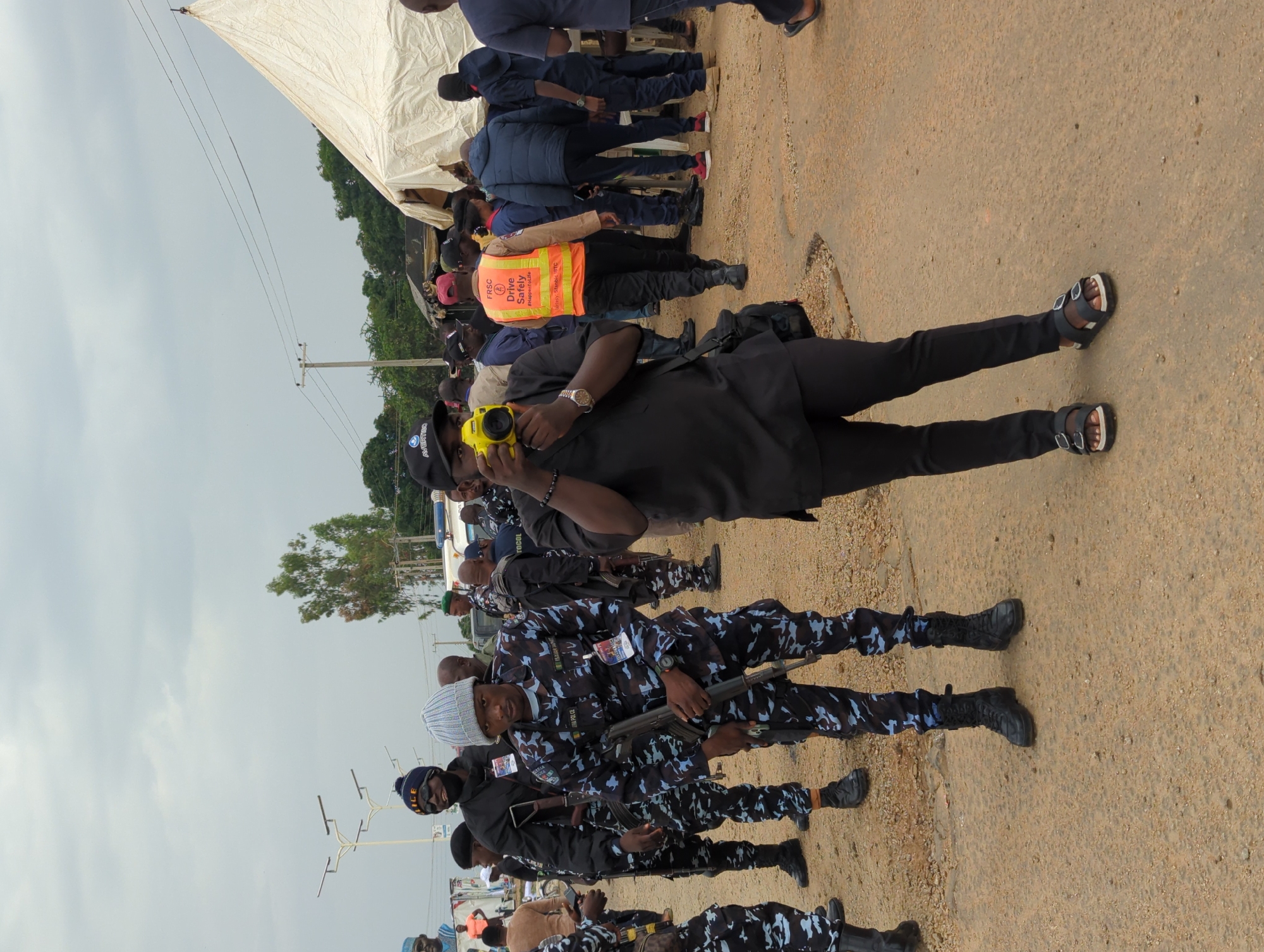 In Maigizo, Nigeria, the image captures a scene on a dirt road where a group of individuals are gathered. A contingent of law enforcement personnel, dressed in dark camouflage uniforms and black boots, stand in formation. One officer, wearing a knitted cap, holds a rifle. To the right, an individual in a black uniform and sandals holds a yellow device to their eye. A man in an orange vest stands among a group of people partially obscured by a white tent in the background. The environment includes an overcast sky and sparse vegetation.
