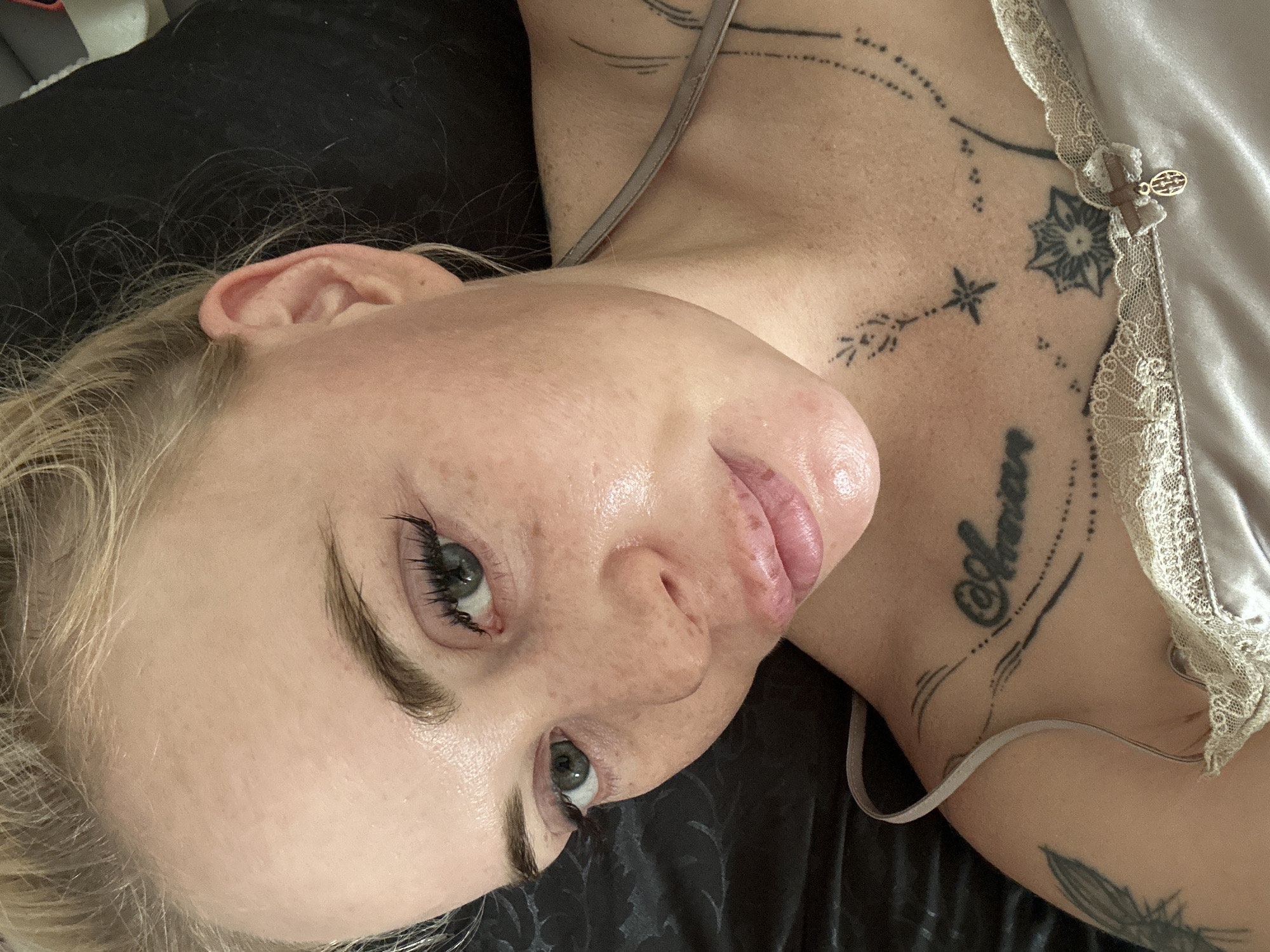A close-up, overhead shot shows a fair-skinned woman with blonde hair, appearing to be in bed, with a black duvet and a silk camisole. She has green eyes, natural eyebrows, and visible freckles across her face. Her lips are slightly parted, and she is wearing mascara and possibly foundation. Tattoos are visible on her chest and shoulder area.
