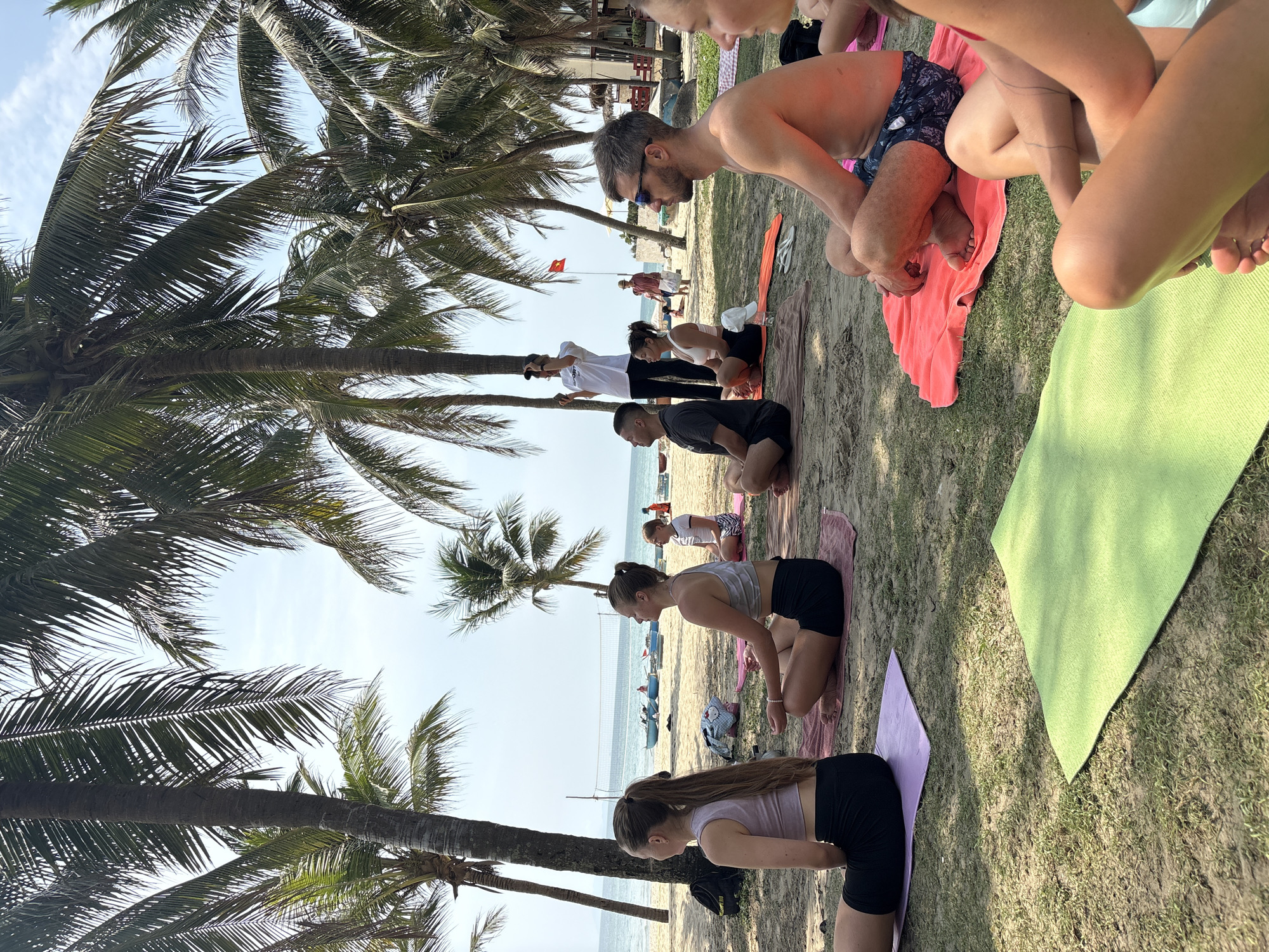 This image captures an outdoor group exercise session, likely yoga or a similar fitness class, taking place on a sunny beach in Đà Nẵng, Vietnam. The scene is set under the shade of numerous tall palm trees, their fronds reaching towards a clear, bright sky.
Approximately eight to ten people, a mix of men and women of varying ages, are visible in the foreground and mid-ground. They are mostly in kneeling or low-to-the-ground poses on individual yoga mats of various colors, including pink, green, and purple. The mats are laid out on a surface that transitions from sandy beach to grassy patches. Some participants are shirtless, while others wear sports tops and shorts. Everyone appears focused on the activity.
In the background, the sandy beach extends towards the ocean, which is visible under a clear sky. Small boats are dotted in the water, and a volleyball net is partially visible further down the shore. A red flag with a yellow star, characteristic of Vietnam, flies from a pole in the mid-ground. Buildings can be seen faintly in the distance. The overall atmosphere is calm and healthy.
The time of day appears to be either morning or late afternoon, indicated by the bright, clear sky and the angle of the sunlight casting shadows from the palm trees and participants. No discernible text is visible on clothing or objects within the image.
