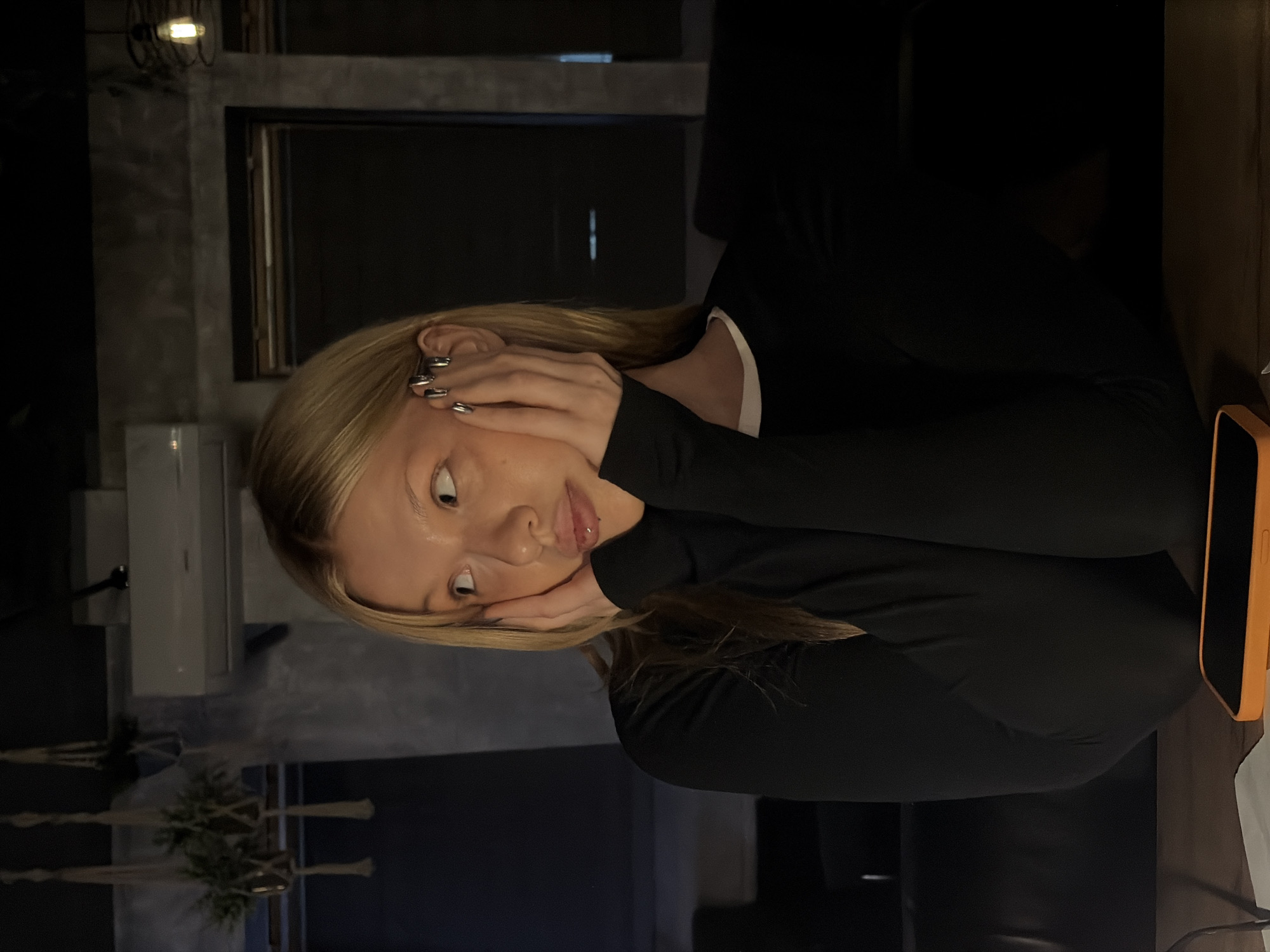 A woman with blonde hair and silver nail polish is sitting at a table in what appears to be an indoor setting. She is wearing a black long-sleeved shirt and resting her face in her hands. An orange phone case is visible on the table to her right. The background includes a grey wall, a black frame with a dark interior, a light fixture, and what looks like a macrame plant hanger. The image was taken in Chaikovsky, Russia.