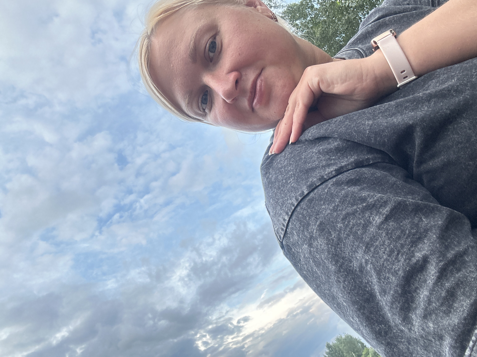 A young, fair-skinned woman with blonde hair is pictured in a close-up, low-angle shot against a cloudy sky. She is wearing a dark gray t-shirt and has her left hand resting on her chin. A light pink smartwatch is visible on her right wrist. The background consists of a sky filled with various cloud formations, with hints of green trees visible on the right side of the frame. The overall lighting suggests it is daytime, possibly late afternoon or early evening due to the diffused light. The location is not identifiable as Chaikovsky, Russia.