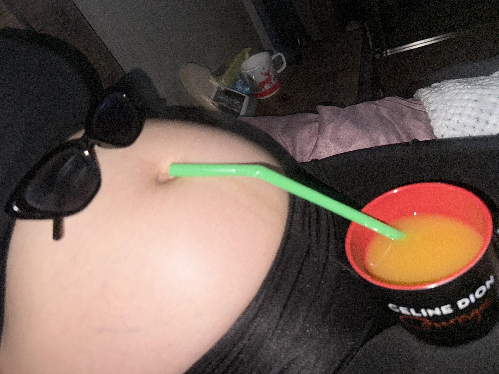 The media file shows a close-up shot of a person's torso, specifically their belly area, with a green straw inserted into their belly button. The person is wearing a black top. To the left of the belly, a pair of black cat-eye sunglasses is visible, perched on the person's stomach. To the right, a black mug with a red rim is filled with an orange liquid, presumably a beverage. The mug has white text on it that reads "CELINE DION Courage". In the background, a wooden surface is visible, along with a Christmas-themed mug and some other blurry objects, suggesting an indoor setting. The lighting appears to be dim, possibly indicating nighttime or a dimly lit room. There are no explicit cues about the weather or specific emotions being conveyed, beyond what might be inferred from the surreal and humorous nature of the setup. The context of Tbilisi, Georgia is provided but not visually evident within the frame.