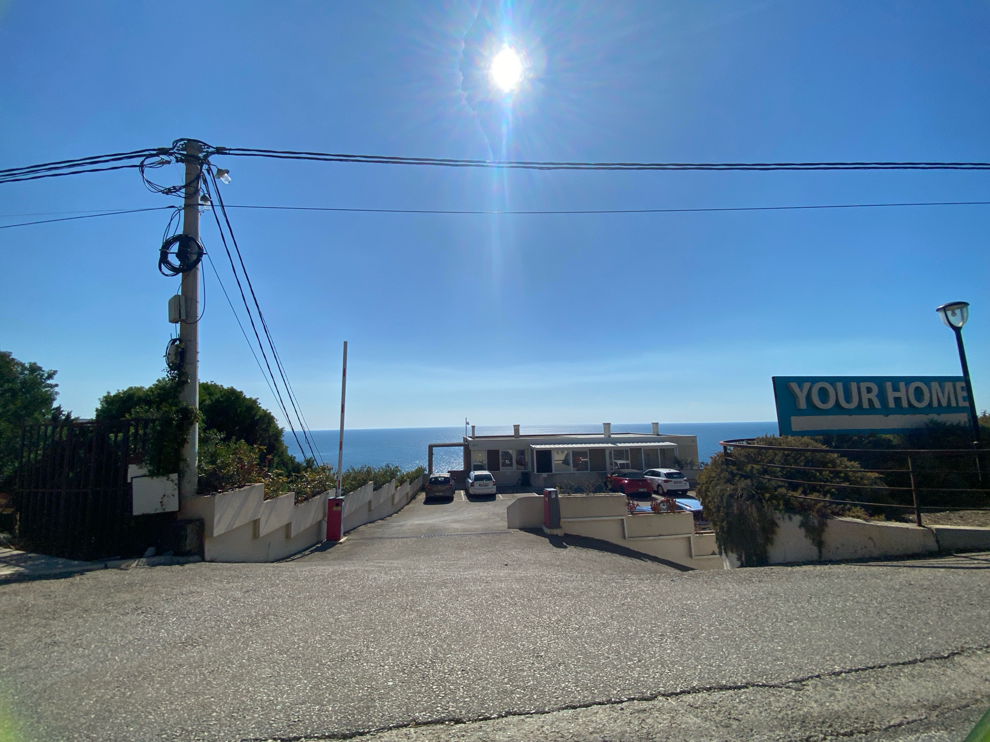 A wide shot captures a sun-drenched scene in Dobra Voda, Montenegro, focusing on a driveway leading towards a building complex situated on a gentle slope overlooking the Adriatic Sea. The sky is a clear, vibrant blue, with the sun positioned high and slightly to the left, creating a bright glare and lens flare. Overhead, power lines stretch across the frame, supported by a utility pole on the left, adorned with numerous cables and some coiled wiring.
The driveway is paved with asphalt, showing some wear and tear. On the left side of the driveway, a black metal fence encloses a property with lush green foliage. A white retaining wall with angled sections separates the driveway from landscaping on both sides. A red automatic gate barrier is positioned near the top of the driveway, with a white pole extending upwards.
Further up the driveway, a building with a flat roof and a glass-fronted section is visible. Several cars are parked in front of it: a dark sedan, a white hatchback, a red sedan, and a white SUV. The building appears to be a commercial establishment or residence, with awnings providing shade over some of the windows. A tall, modern street lamp stands to the right of the building, and further to the right, a large blue sign with white lettering reads "YOUR HOME." The sea stretches out to the horizon, calm and reflective under the bright sun. The overall impression is one of a sunny coastal location.