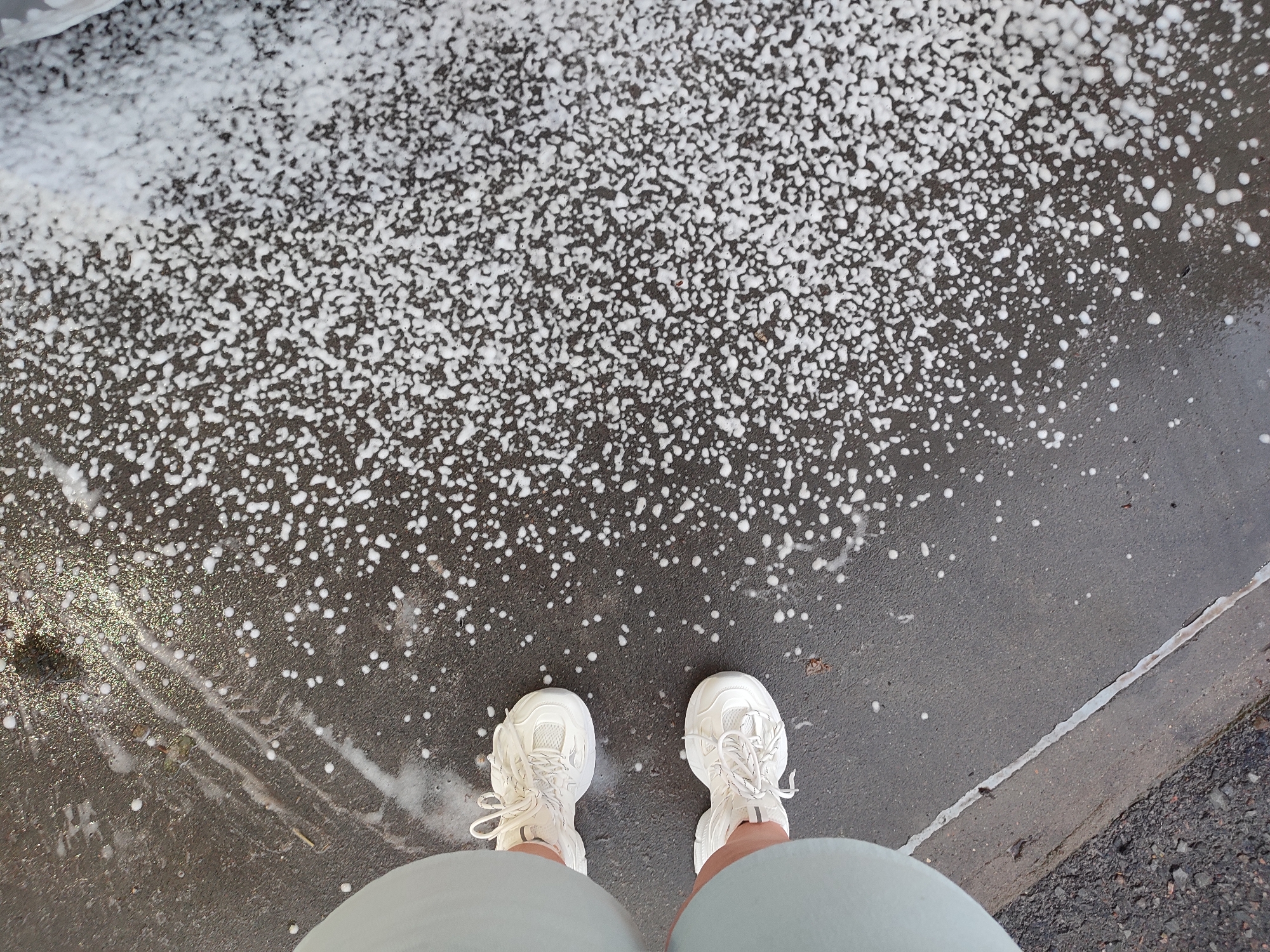An overhead, eye-level perspective shot captures a pair of white lace-up sneakers on a wet, dark asphalt surface. The sneakers are positioned slightly apart, facing forward. The ground is covered in scattered white suds and water droplets, with a distinct line of foam running diagonally from the top left of the frame towards the bottom right. A sidewalk curb is visible on the right, transitioning to a gravelly surface. The overall scene suggests a car wash or an area recently cleaned with soap and water. The location is identified as Voronezh, Russia.