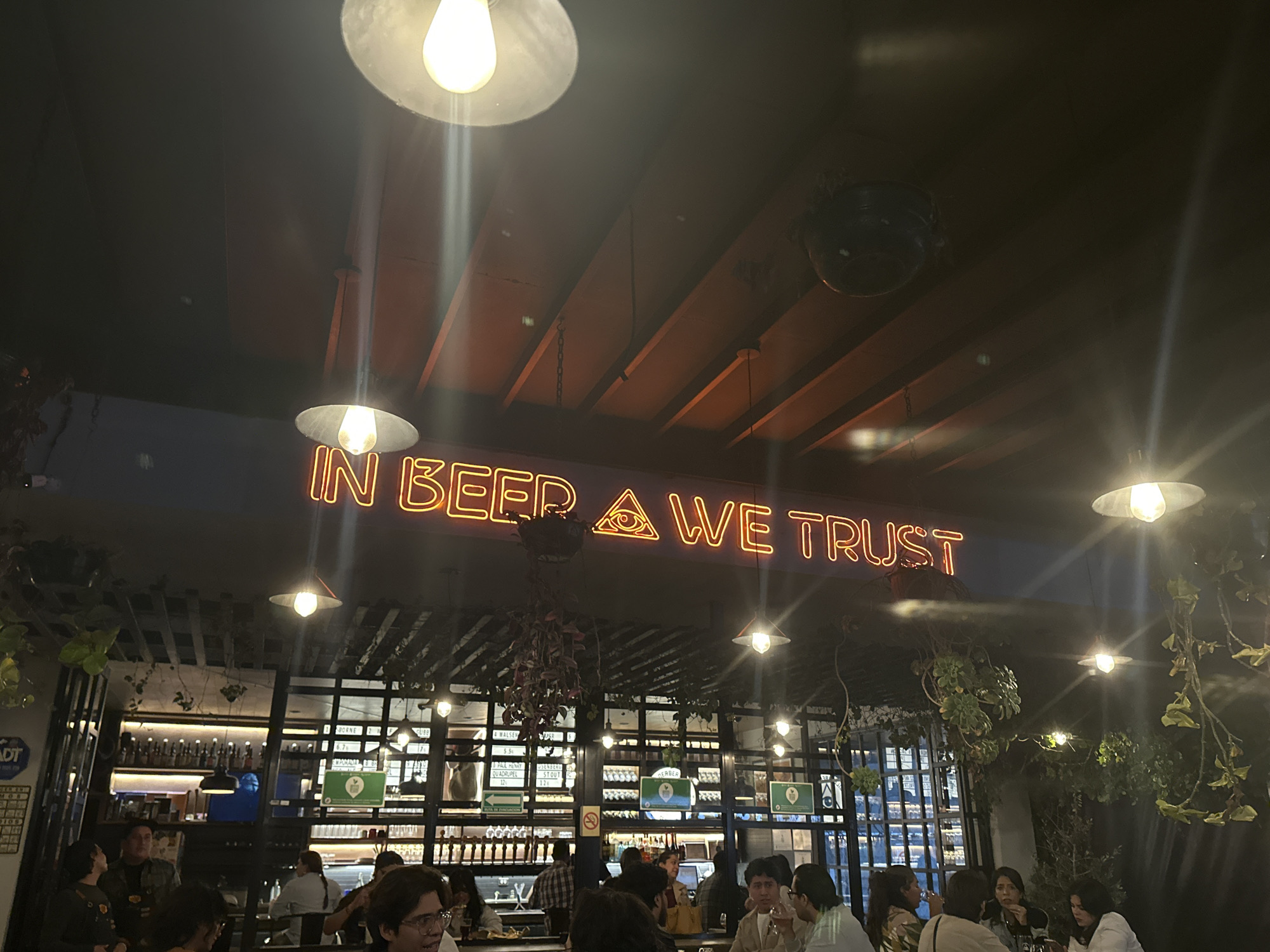 Captured inside a dimly lit establishment in San Andrés Cholula, Mexico, a diverse group of individuals is seated at tables, engaging in conversation and activity. The space features a bar with shelves stocked with bottles and a menu of various beverages listed on the wall. Neon signage prominently displays the phrase "IN BEER WE TRUST," with an eye symbol. The environment is adorned with hanging plants and exposed beams in the ceiling.