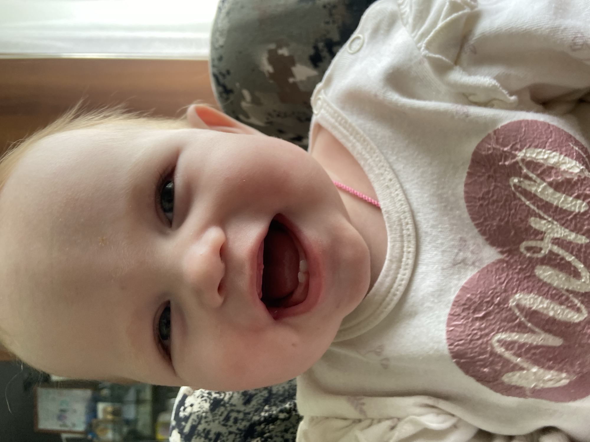 A close-up, slightly angled shot shows a baby with fair skin and light blond hair lying down. The baby's mouth is open, revealing several teeth and the inside of its mouth. The baby is wearing a white shirt with a pink design on it. A pink necklace can be seen around the baby's neck. The background includes furniture with a patterned design. Based on the provided context, the location is specified as Фоки, Russia.