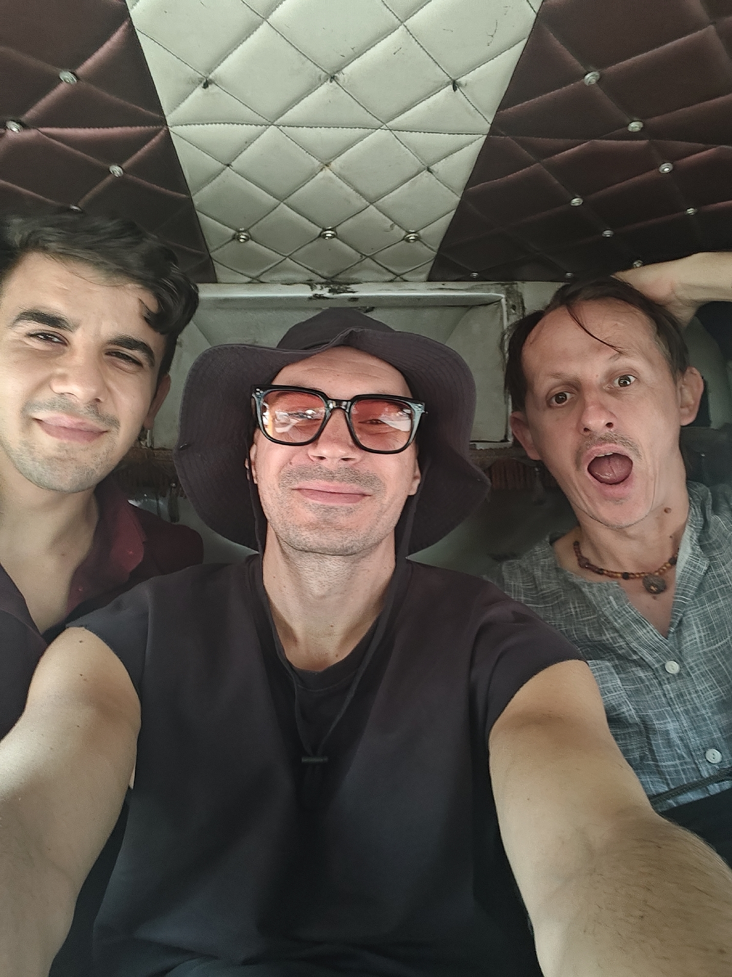 A close-up, front-facing selfie photograph shows three adult males seated together inside a vehicle in Unknown, Unknown. The man on the left, with dark hair and a light beard, wears a dark red collared shirt and smiles subtly. The central figure, taking the photo with an extended arm, wears a dark wide-brimmed hat, black-framed sunglasses with orange-tinted lenses, a dark sleeveless shirt, and a black cord necklace; he also smiles faintly. The man on the right, with light brown hair and a light mustache, wears a light blue/grey textured button-up shirt and a beaded necklace with a circular pendant. He has a wide-eyed, open-mouthed expression, and his right hand is positioned behind his head. The background reveals the vehicle's ceiling, which is upholstered in a diamond-quilted pattern, divided vertically into a dark maroon section on the left and an off-white section on the right, both adorned with reflective studs at the intersections. The off-white section shows visible dirt and wear, and a white structural element is present below it on the right.