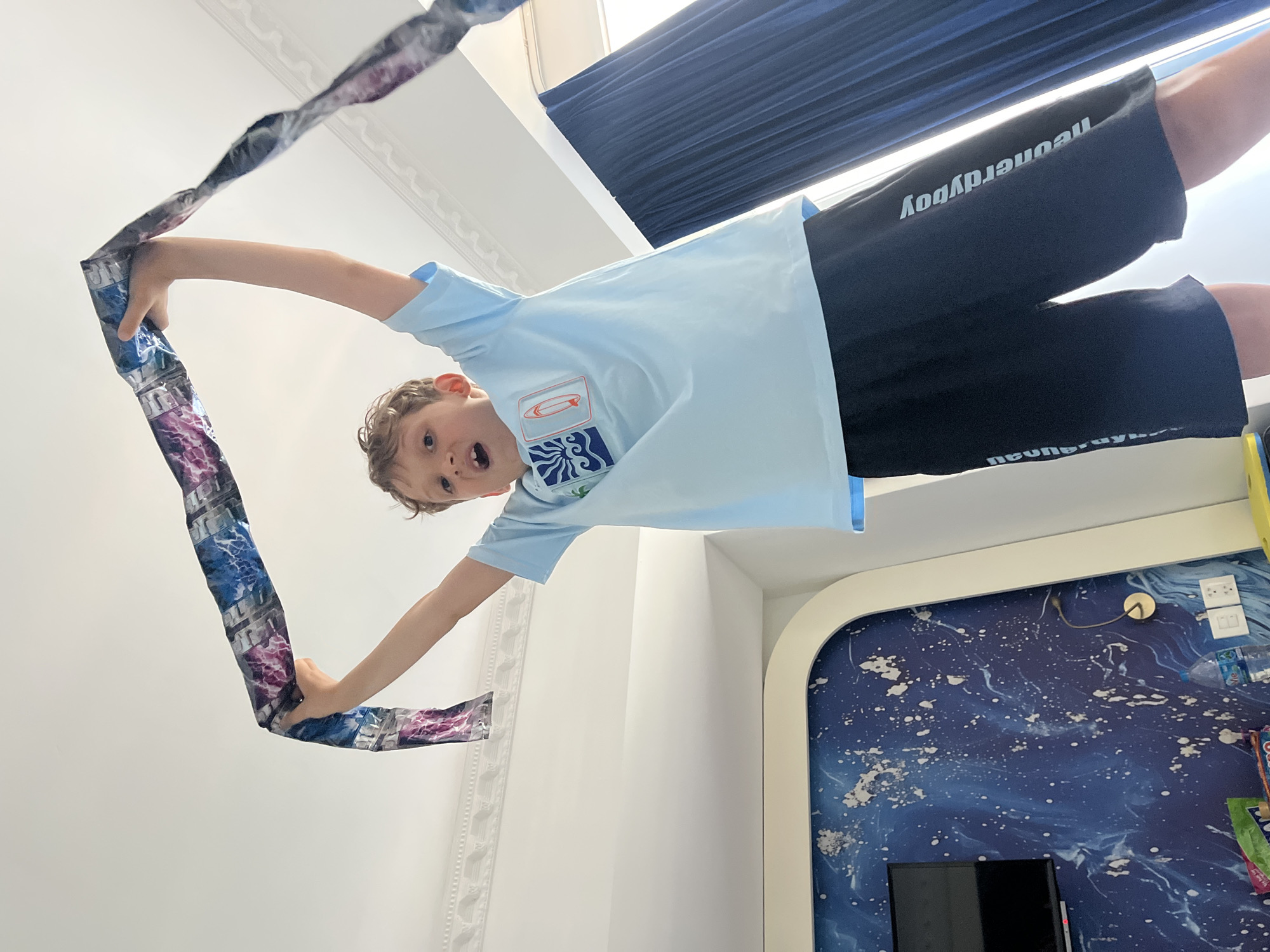 A young boy with curly brown hair stands indoors, holding a long, colorful scarf or piece of fabric above his head. He is wearing a light blue t-shirt with a graphic design and black shorts that have white lettering down the sides. The boy is looking up and appears to be excited or surprised. The background shows a white ceiling with decorative molding, a dark blue curtain, and a wall with a dark blue abstract design. A portion of a television screen is visible on the wall. The overall setting suggests an indoor residential or hotel environment.