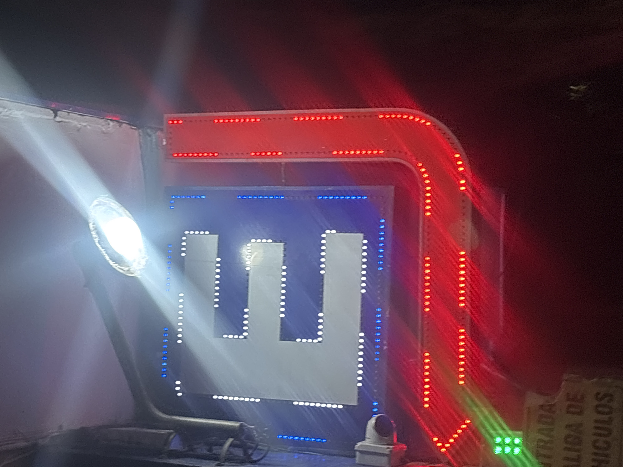 A brightly illuminated sign, predominantly red, white, and blue, is viewed at night. The sign contains a white "E" shape formed by white LED lights on a blue background. The sign is framed by red LED lights forming an arrow shape, pointing downward. A bright spotlight is shining on the left side of the sign. The lower right corner of the image shows part of a sign with text, "ENTRADA" and "SALIDA DE VEHICULOS" suggesting the location is a parking area. Based on the Spanish language and architectural style, the location is likely in Buenos Aires, Argentina. There is a small white box at the base of the sign.