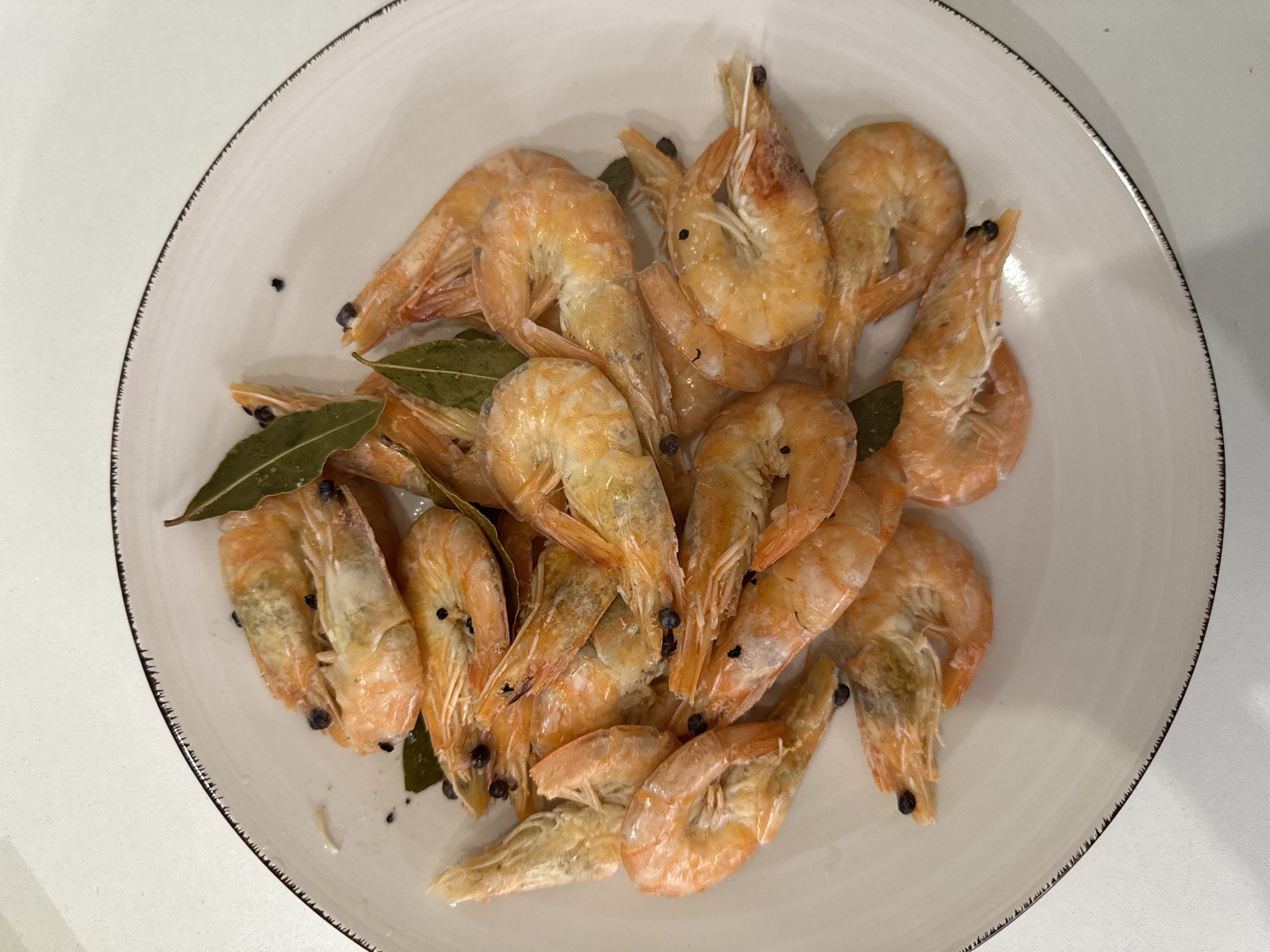 A white, round plate filled with cooked shrimp, bay leaves, and peppercorns sits on a white, speckled countertop. The shrimp are pinkish-orange and curled. Several green bay leaves are interspersed among the shrimp, and small, dark peppercorns are visible throughout the dish. The plate has a dark, jagged trim. The scene appears to be indoors, likely a kitchen. There is no indication of the city of Vologda, Russia.