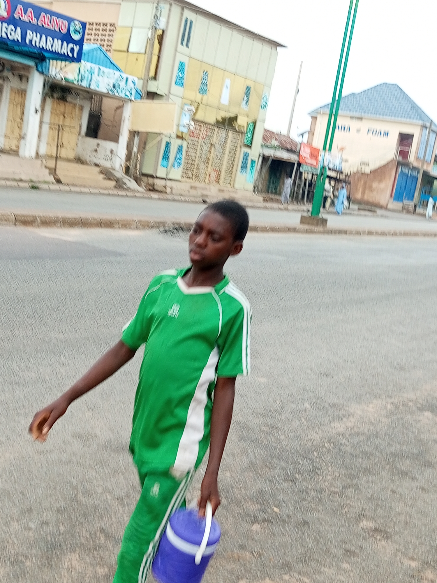 A young person wearing a green tracksuit with white stripes is walking on a paved road in the city of Gombe, Nigeria, carrying a blue bucket with a white handle in their right hand. The person's head is turned to the side, and their eyes are closed or looking down. In the background, there are several buildings, including a pharmacy with blue and white signage, and other commercial establishments. The sky is overcast. Several other people are visible in the background, walking on the sidewalk or near buildings. A green pole with multiple vertical supports stands prominently in the mid-ground. The road appears to be a commercial street.