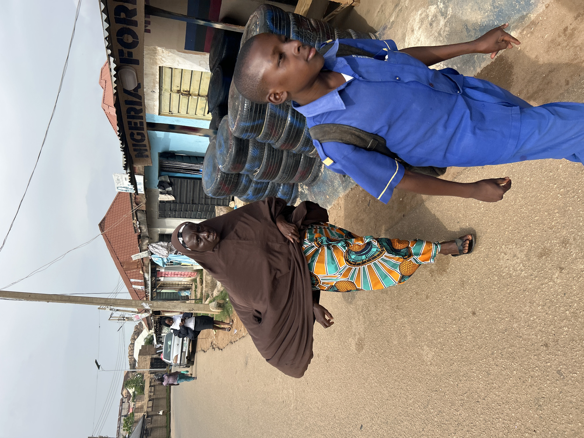 In Jos, Nigeria, a brown-robed woman with a patterned skirt walks down a paved street. Beside her, a boy in a blue uniform with a backpack walks forward. A stack of tires is visible on the right side of the image, near a storefront with a "Nigeria For" sign. Other people and vehicles are further down the street.