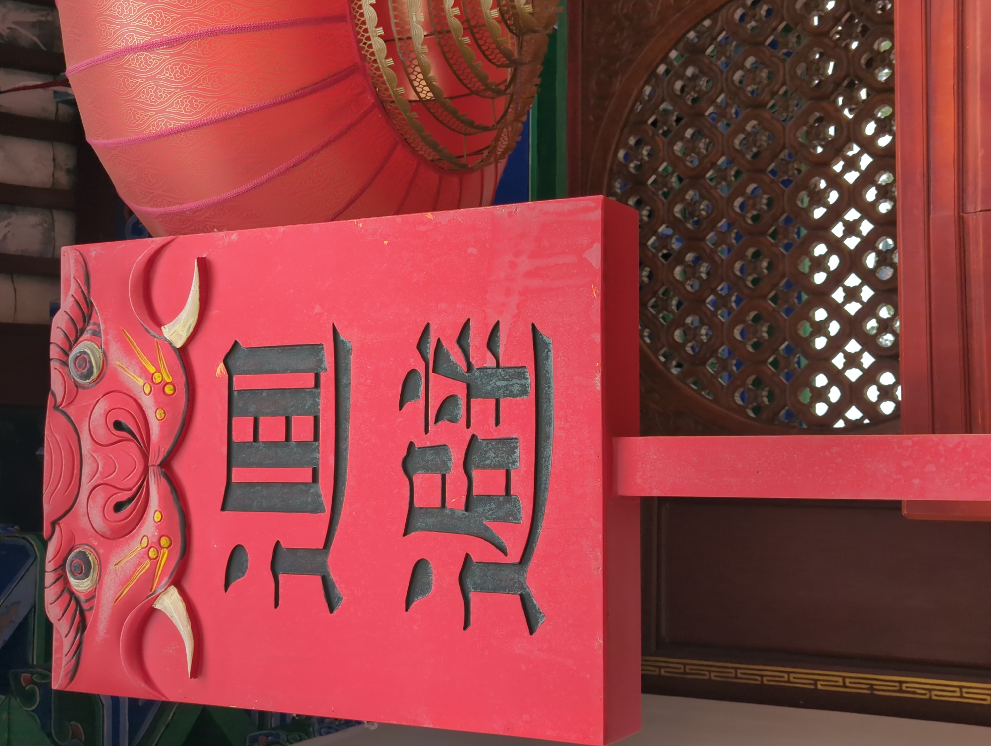 This is a photograph of a red wooden sign with black Chinese characters carved into it. The sign is adorned with a red, dragon-like motif on the left side, featuring carved eyes and a horn. The Chinese characters appear to read "景觀" (jǐngguān), meaning "scenery" or "view."
Behind the sign, a large, ornate red lantern with gold embellishments hangs from above. To the right of the sign, a wooden lattice window with intricate geometric patterns and colored glass inserts is visible. The overall setting suggests a traditional Chinese architectural environment, possibly a temple, garden, or a historic building. The red color of the sign and lantern, combined with the gold and intricate designs, evokes a sense of cultural richness and celebration. The lighting suggests it is daytime, with natural light illuminating the scene. The photograph is taken at a close-up angle, focusing on the sign and its immediate surroundings. There is no visible indication of people or a specific event taking place, but the elements present suggest a place of cultural significance or historical interest in Gucheng District, China.