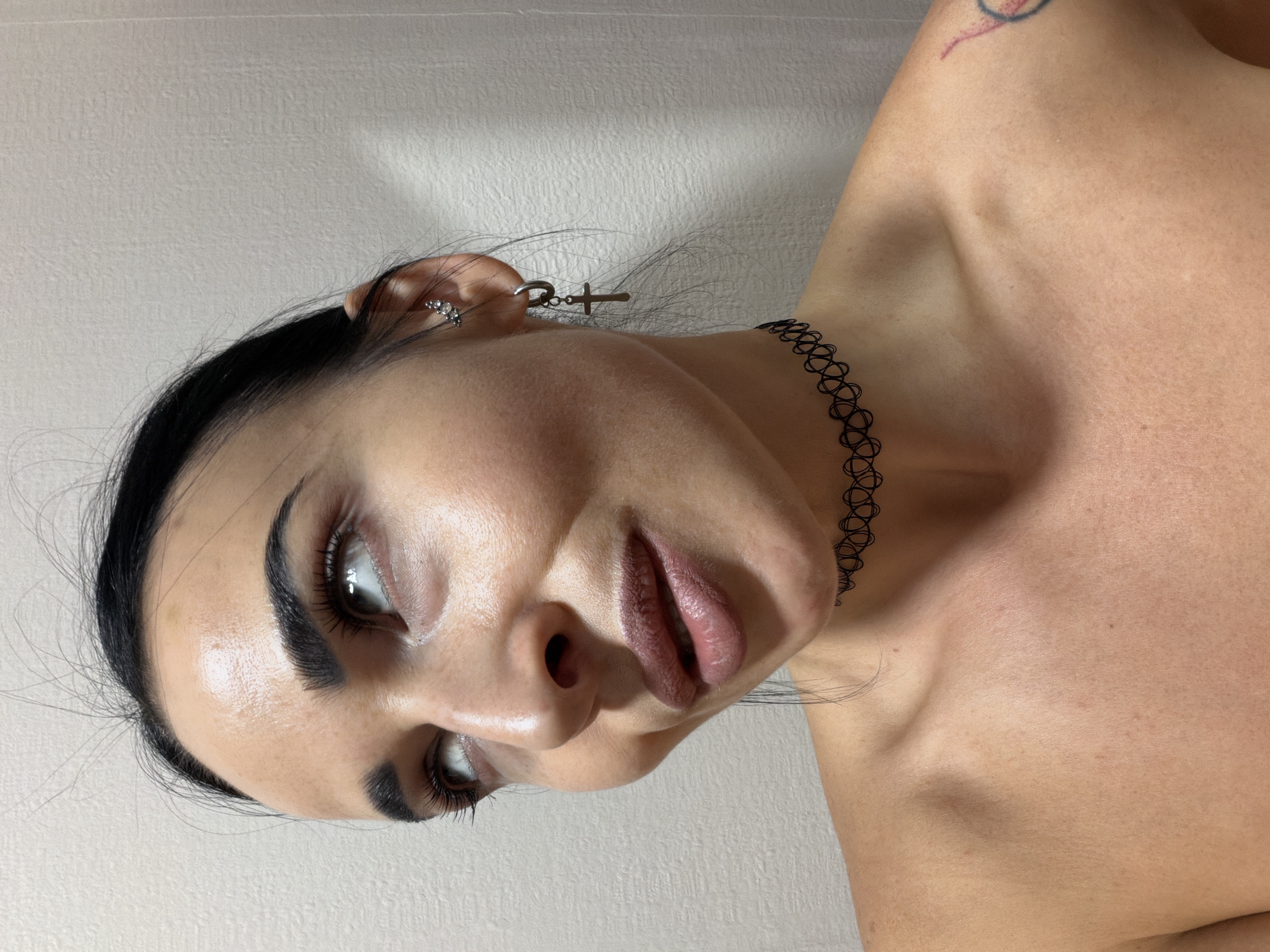 A close-up portrait features a person, likely female, with dark hair and prominent facial features. The image is oriented sideways, with the subject's head tilted and their gaze directed slightly upwards and to the left.
The subject has strong, dark eyebrows, pronounced eyeliner, and mascara defining their eyes. Their lips are full and appear to have a muted pink or nude lipstick. The skin on their face and visible bare shoulders/chest is fair, showing natural texture and some reflective highlights, particularly on the forehead and nose.
Around their neck, they wear a black, open-work, elastic choker necklace. The visible ear features multiple silver piercings: a cluster of small, sparkling studs higher up on the ear and a dangling earring with a small, silver cross shape. A small, stylized or abstract tattoo in faded red and blue ink is visible on the upper right arm/shoulder area.
The background is a plain, light-colored wall with a subtle textured finish. The lighting appears soft and even, suggesting an indoor setting. There is no discernible activity other than posing for the portrait. The subject's expression is neutral or contemplative. No text is visible in the image.