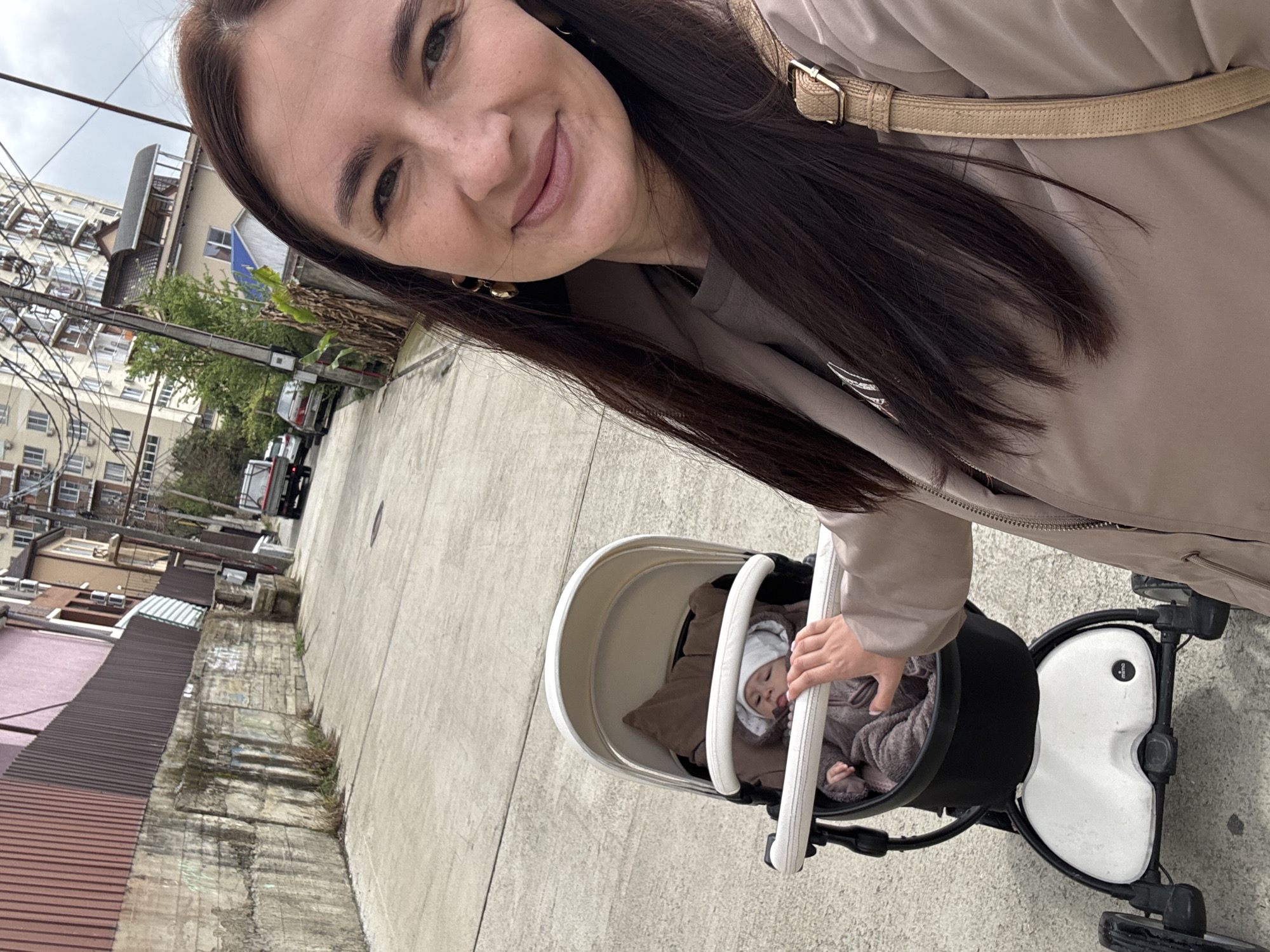 The image shows a woman holding a stroller with a baby in it. The woman is fair-skinned with dark hair and is wearing a beige jacket with a strap across her chest. The baby is wearing a grey patterned outfit and a white bonnet. They are on a sidewalk in what appears to be a city. Buildings line the street with an overcast sky in the background. The location is Sochi, Russia.