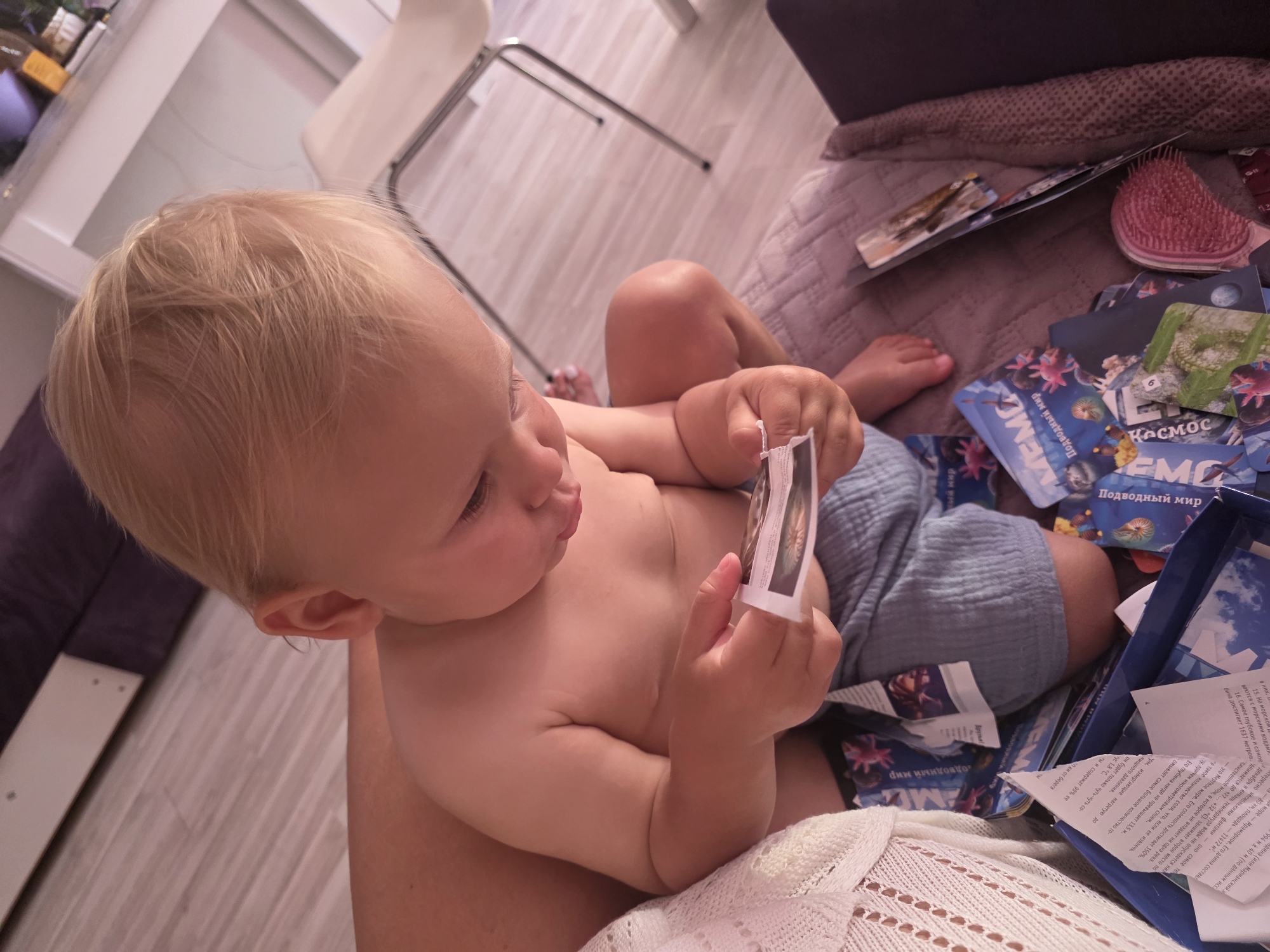 In a brightly lit indoor setting, a baby with fair hair and rosy cheeks is seated on a blanket. The baby is wearing light blue shorts and is holding a small card in their hands, examining it with curiosity. Around the baby, a collection of cards, likely from a memory or educational game, are scattered. The cards feature images and text related to "Космос" (Space) and "Подводный мир" (Underwater world), with one card clearly showing the word "MEMO." The scene appears to be set in a home in Sochi, Russia, with wooden flooring visible. The overall atmosphere is one of a child engaged in play and learning.