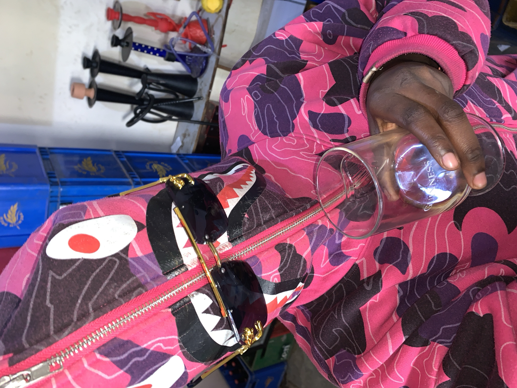 A person, whose face is obscured by a pink and purple camouflage hooded jacket with a shark mouth graphic and gold-rimmed sunglasses, holds a clear glass in their right hand. The jacket has a prominent zipper and is adorned with a stylized shark face. In the background, stacks of blue crates are visible, along with several hookah pipes and a yellow object on a shelf. The scene appears to be indoors, possibly in a shop or recreational area in New Stadium, Nigeria. The glass has the word "GUINNESS" etched on it.