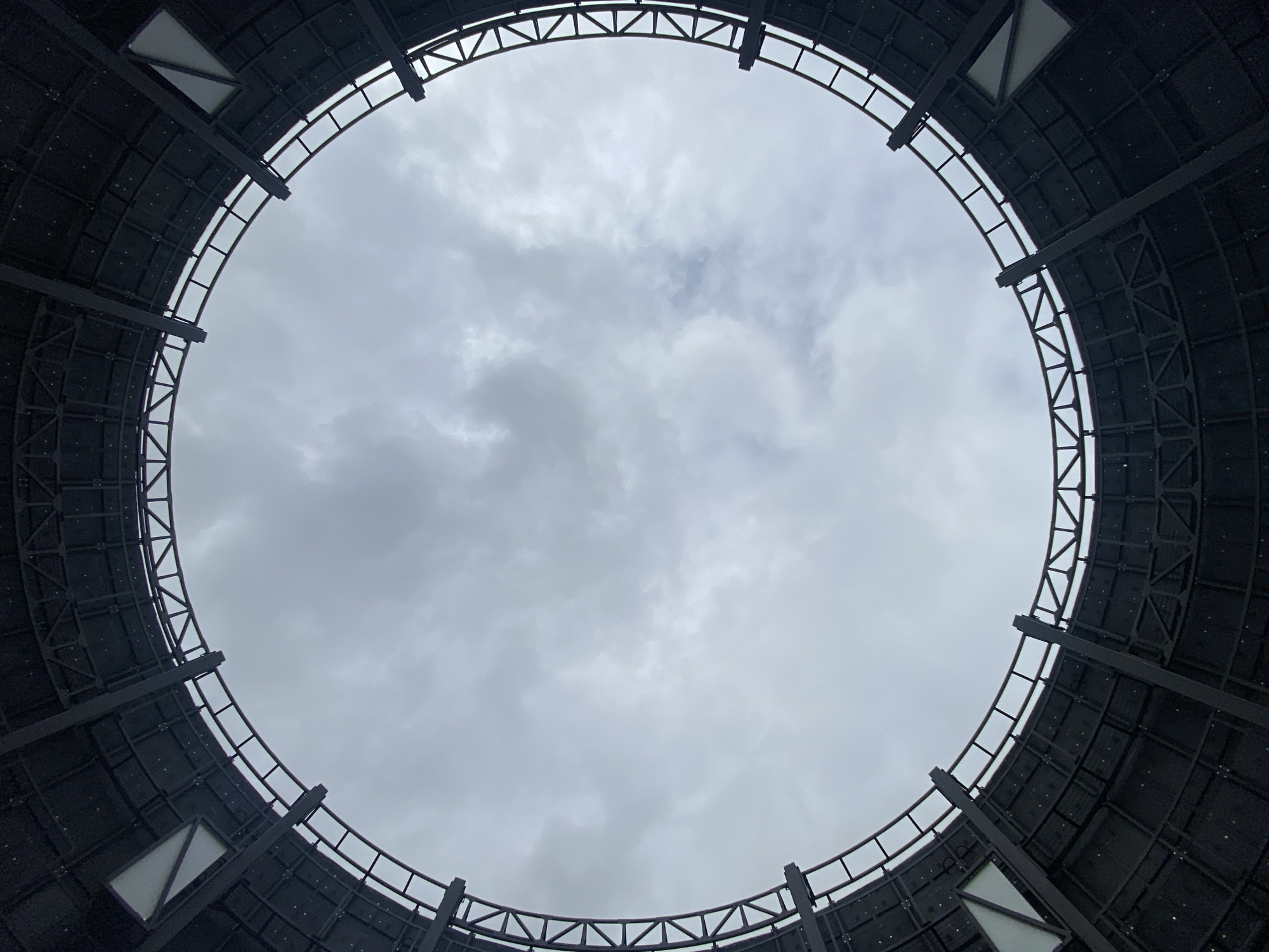 The image shows a view looking up through the center of a large, circular structure in Zavolzhye, Russia. The structure has a steel framework with panels filling in the spaces between the frame. The sky above is overcast and filled with gray clouds. Several white triangle-shaped structures are visible within the frame on two sides, possibly windows. No people are visible.