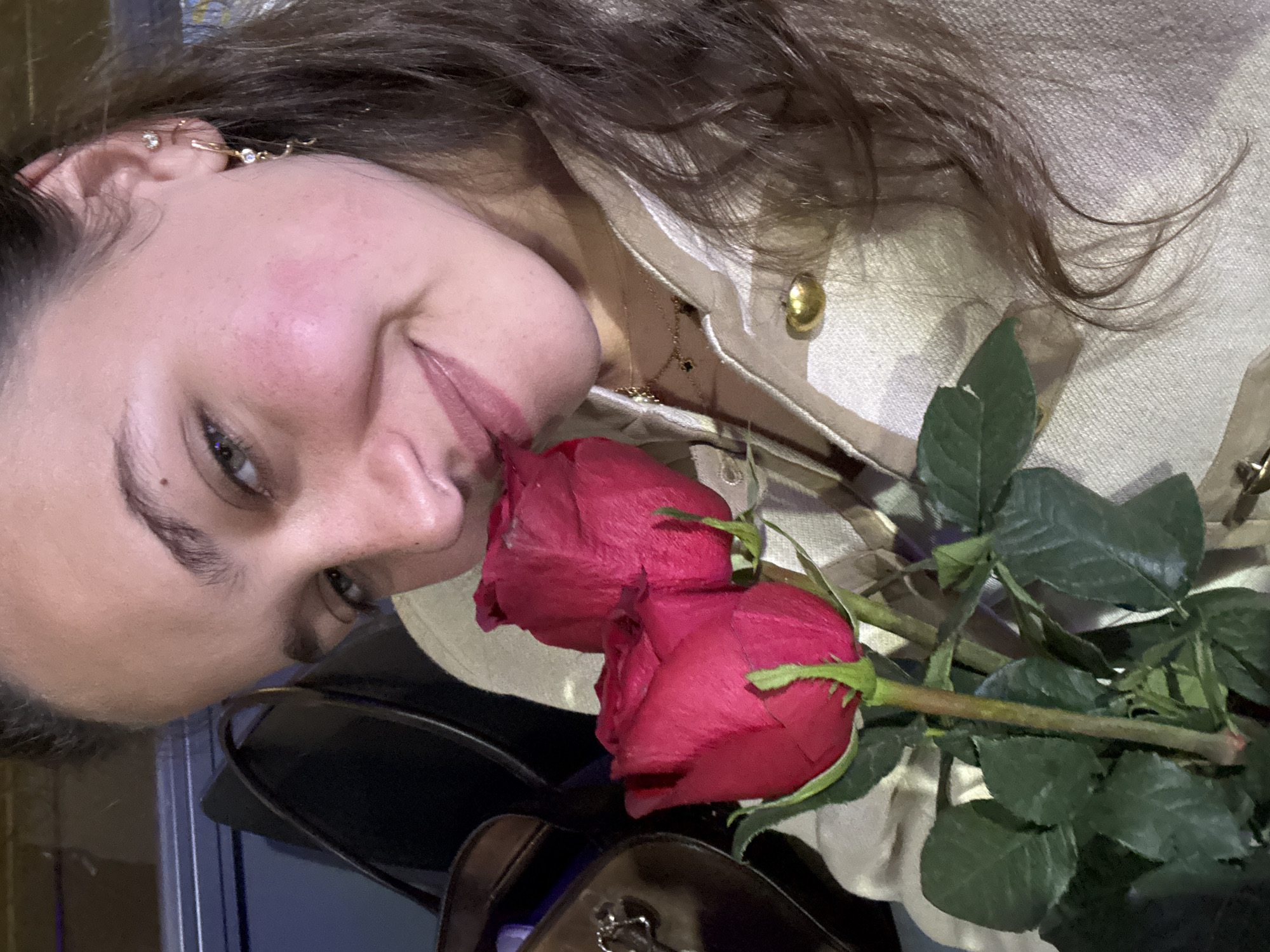 A woman with fair skin, brown hair, and green eyes is seen in a close-up portrait, positioned at an approximate 45-degree angle facing the camera. She is smiling slightly. She is wearing a beige textured jacket with a gold button. A delicate gold necklace is visible. She has multiple gold earrings in her left ear. A bouquet of two red roses is positioned near her face, partially obscuring the lower part of her face. The background includes a blurred dark surface and a glimpse of blue, possibly indicative of an indoor environment. Given the limited details, it's impossible to determine the location as Milan, Italy. There is no concrete information to confirm the city.
